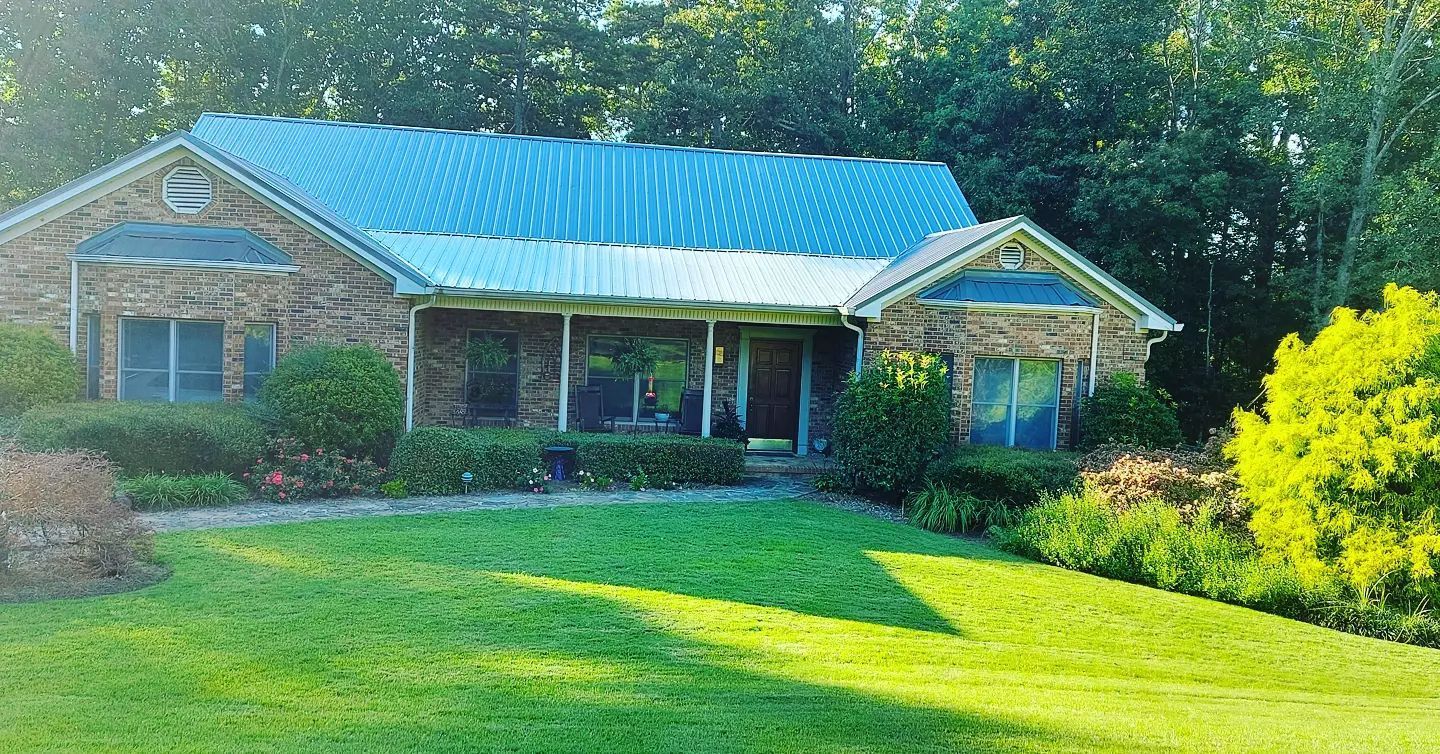 Single-story house with blue metal roof, front porch, and bright green lawn in sunny yard