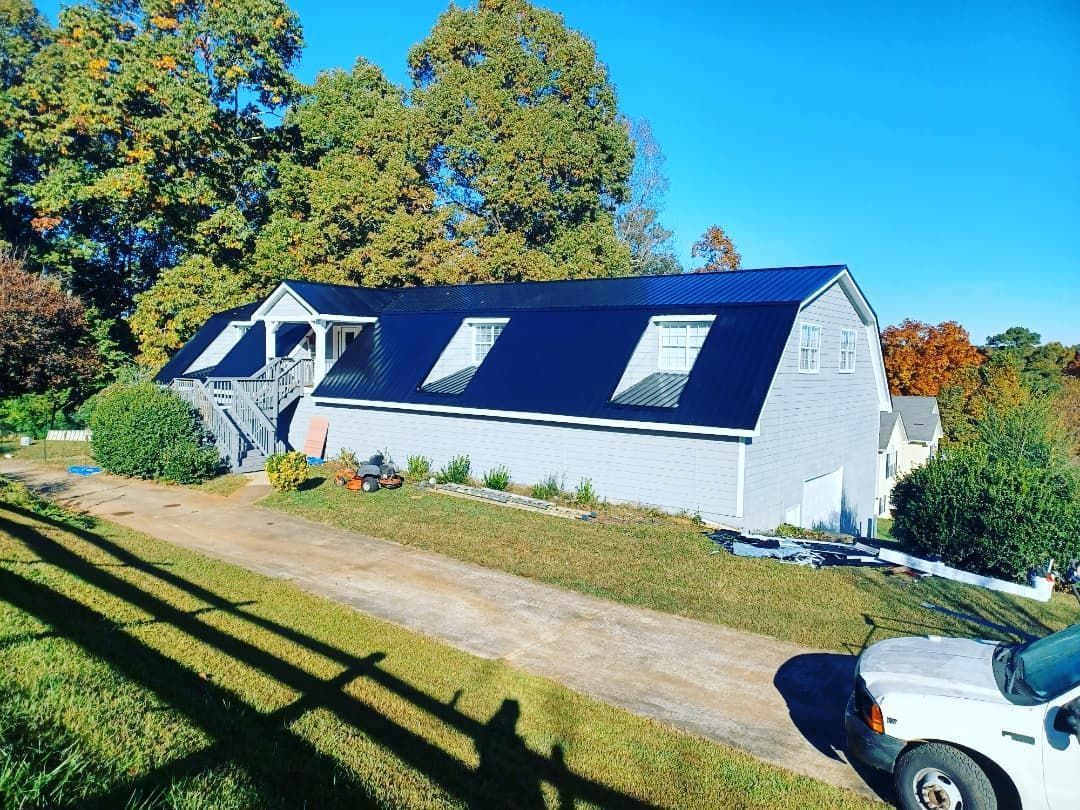 White house with dark roof and skylights on a sunny hillside, trees behind and a car at the right edge.