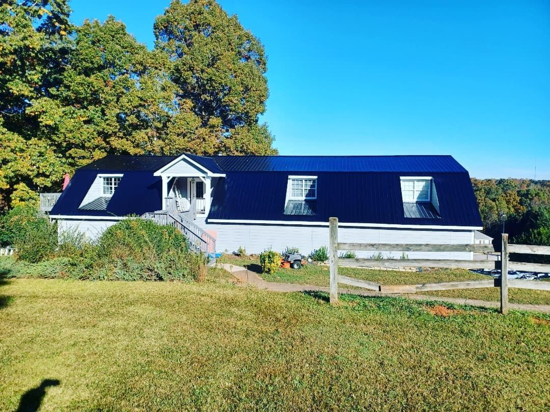 White house with dark roof and dormer windows in a sunny rural field, with trees and a fence nearby