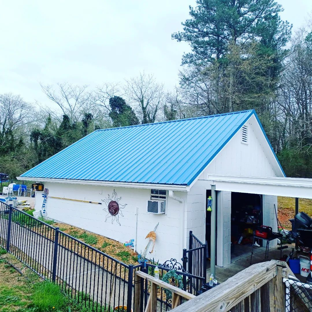 Small white house with a blue metal roof beside a fenced yard and trees.