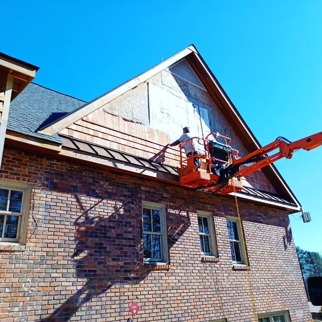 Worker on an orange lift painting a brick house’s upper gable under a blue sky