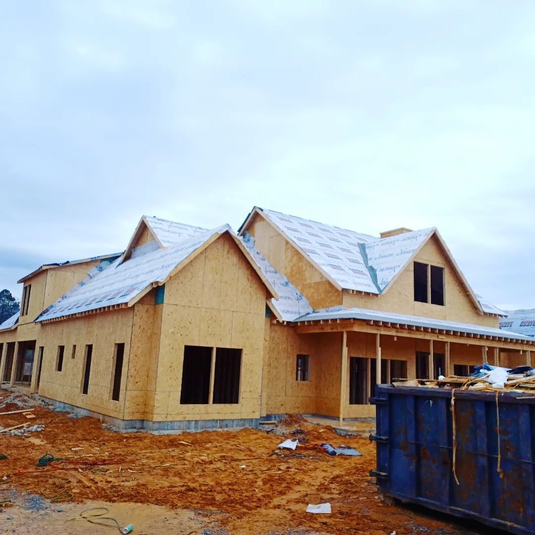Unfinished tan house with gray roof under construction on a muddy lot, with a blue dumpster in front