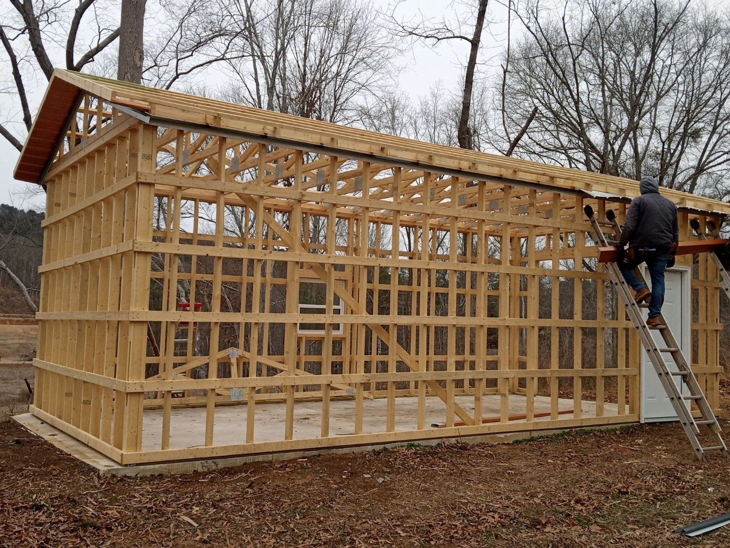 Wooden shed frame under construction, with a worker on a ladder using a power tool.