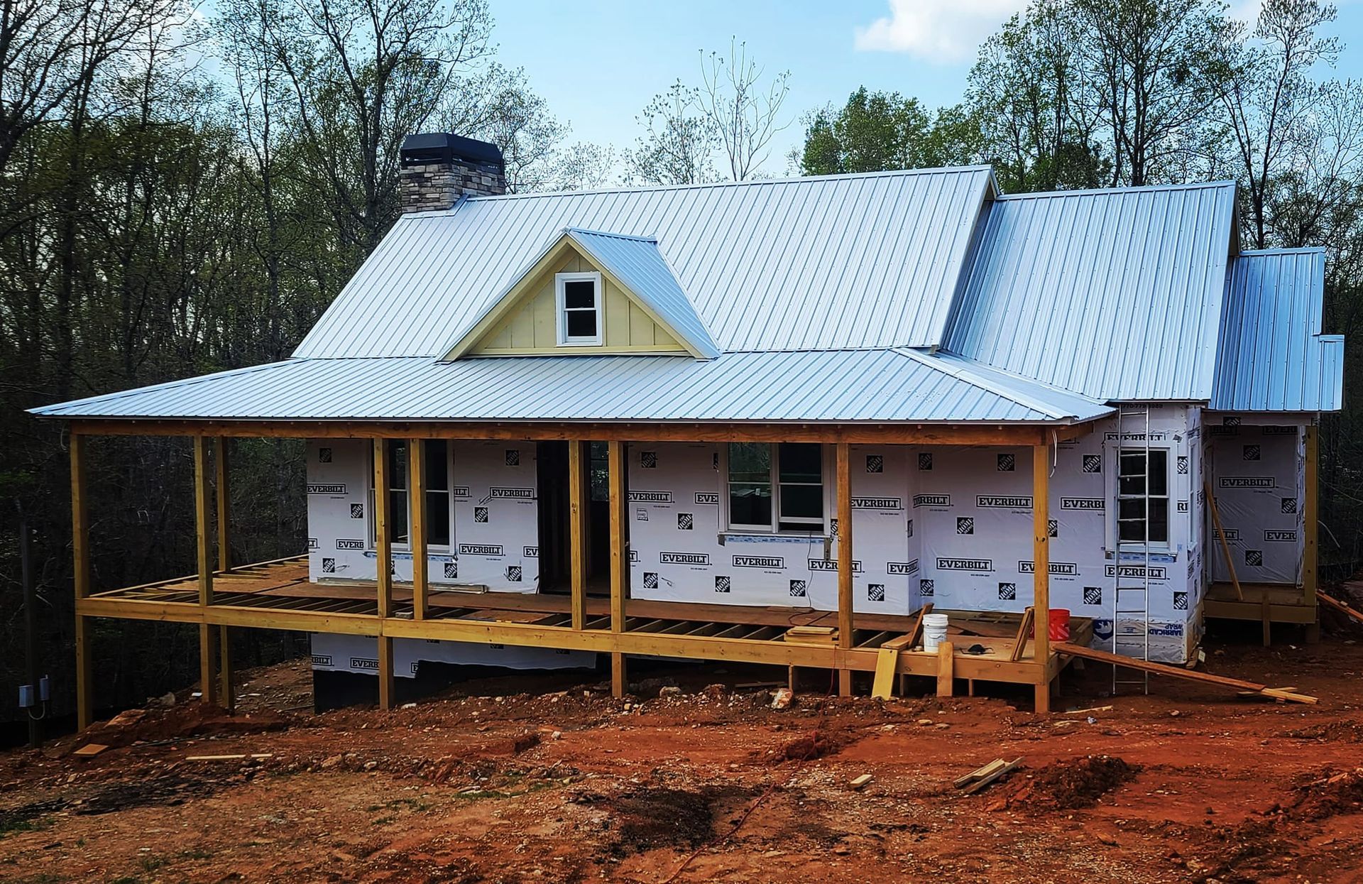 Partially built house with white roof and yellow porch in a wooded, muddy clearing