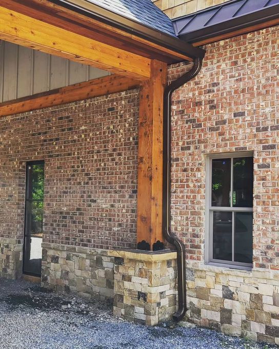 Brick house exterior with timber porch posts, stone base, and a window under a covered roof.