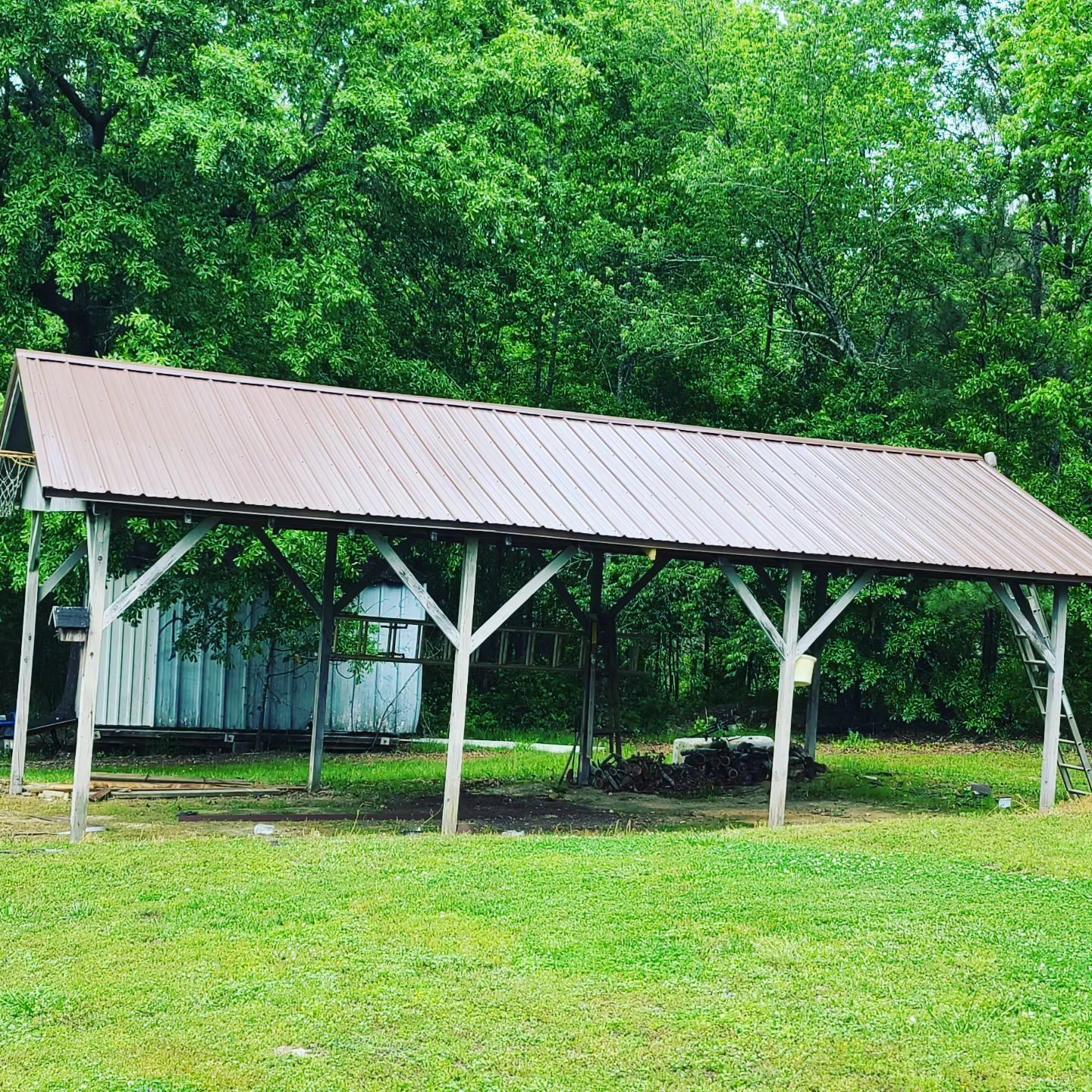 Wooden pavilion with a metal roof in a green park, surrounded by trees and grass.