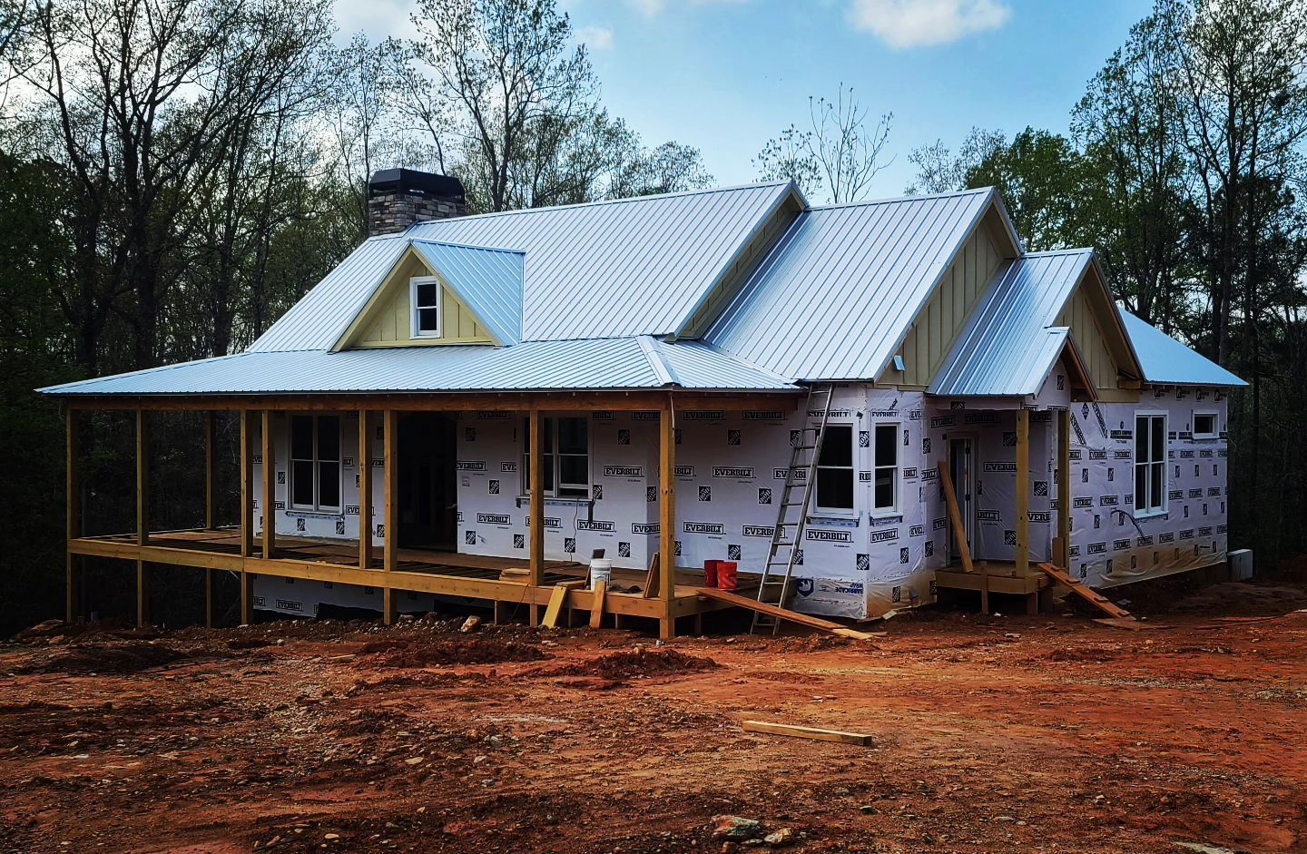 Partially built white house with a metal roof and wraparound porch on a cleared wooded lot