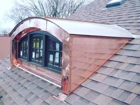 Roof dormer with arched black French doors and copper siding on a shingled house roof.
