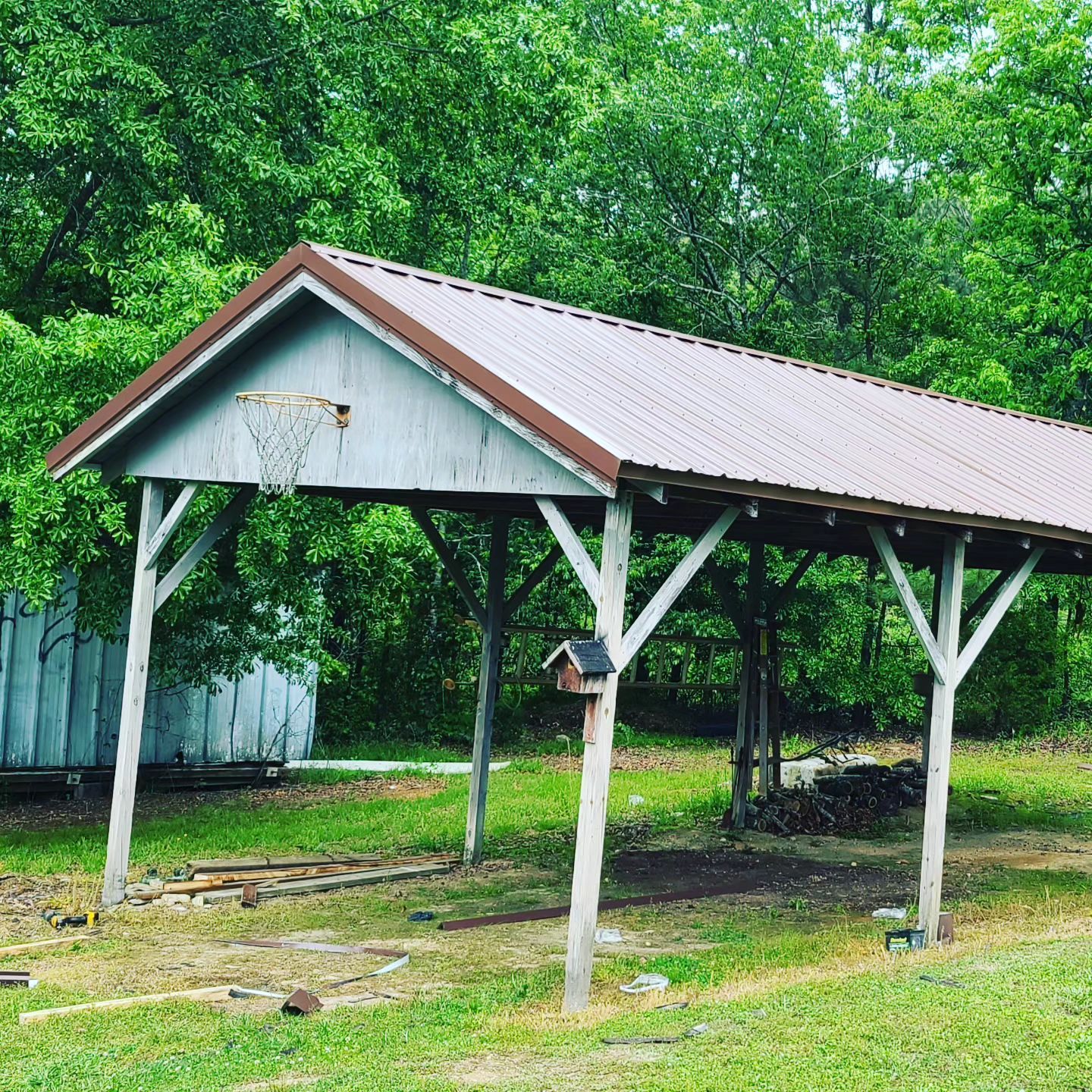 Open-sided shed with a red roof in a green yard, surrounded by trees and grass