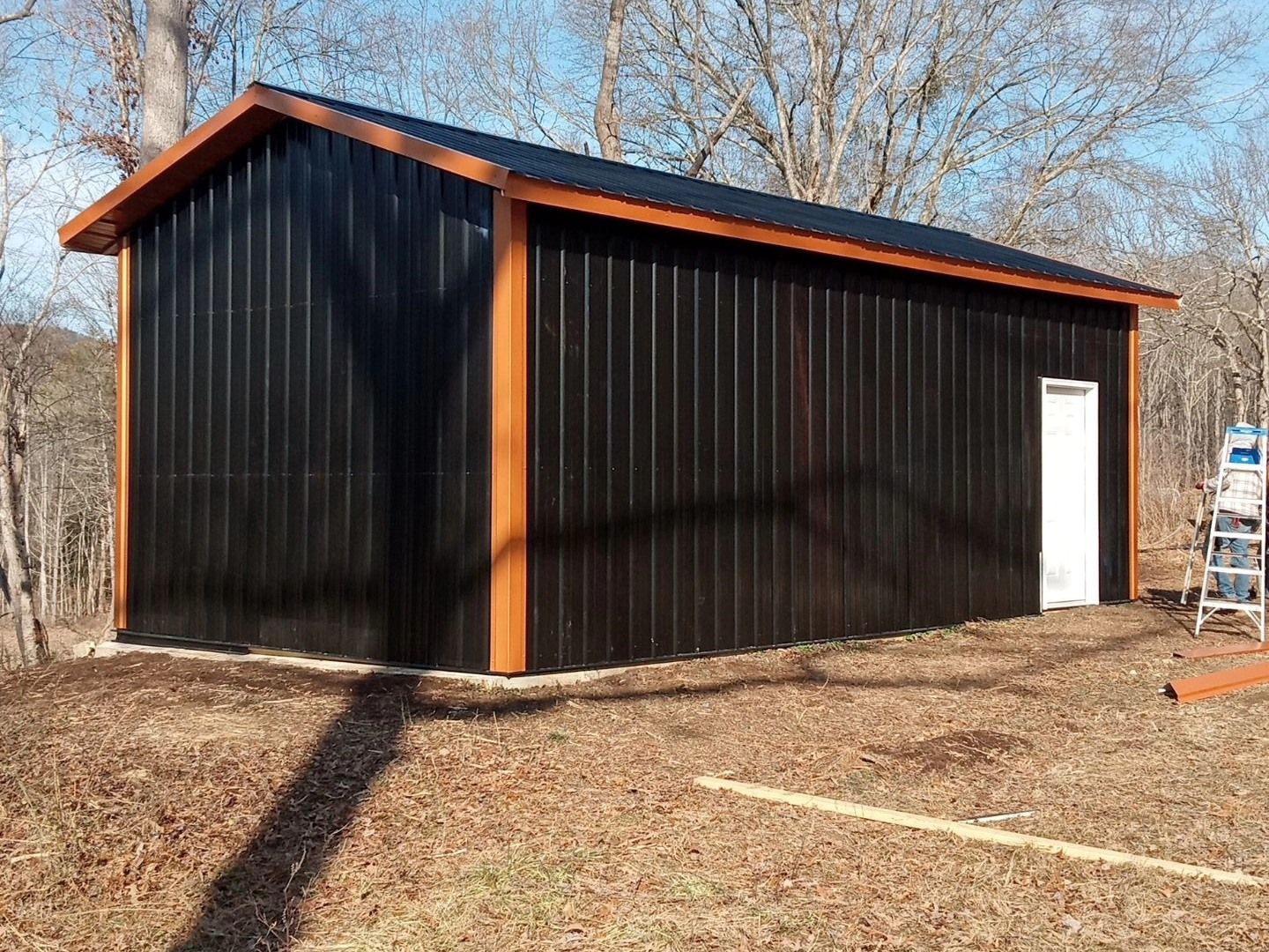 Black metal shed with orange trim and a white door on a wooded lot.