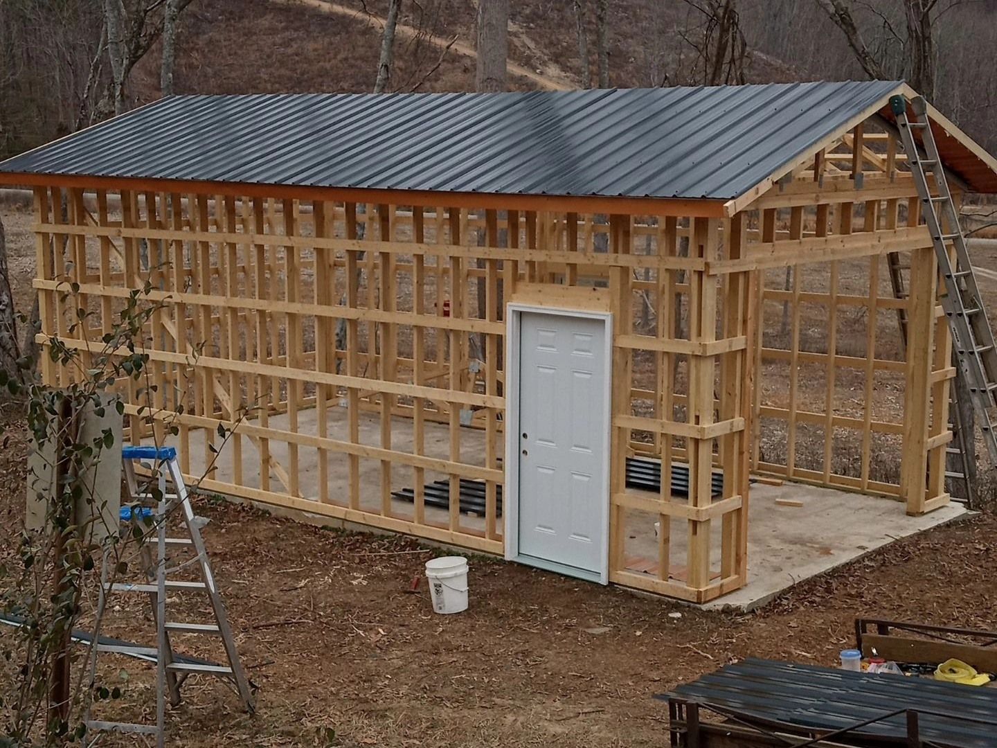 Wood-framed shed under construction with a metal roof, white door, and open dirt surroundings