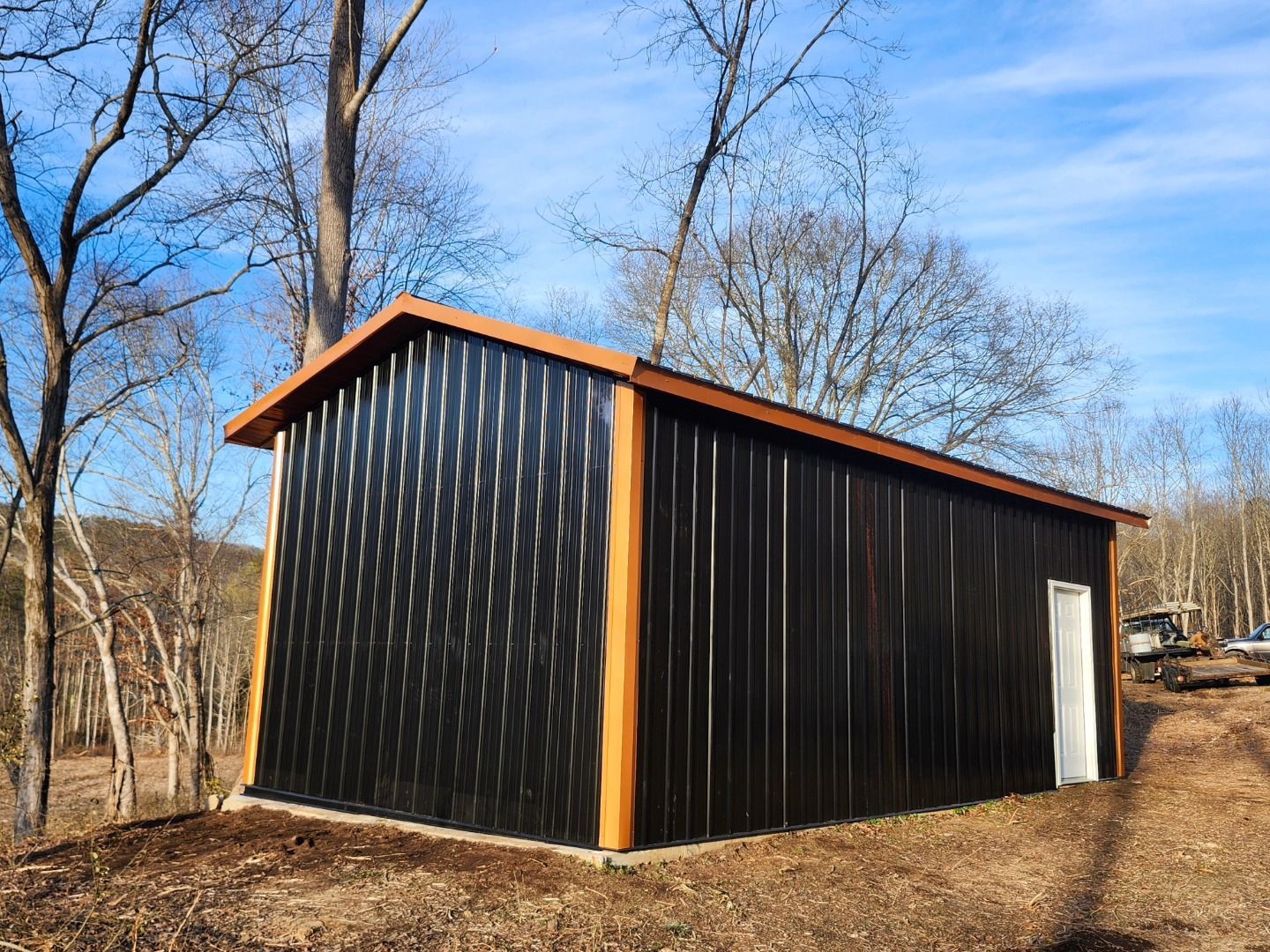 Black metal shed with orange trim in a wooded outdoor area under a blue sky