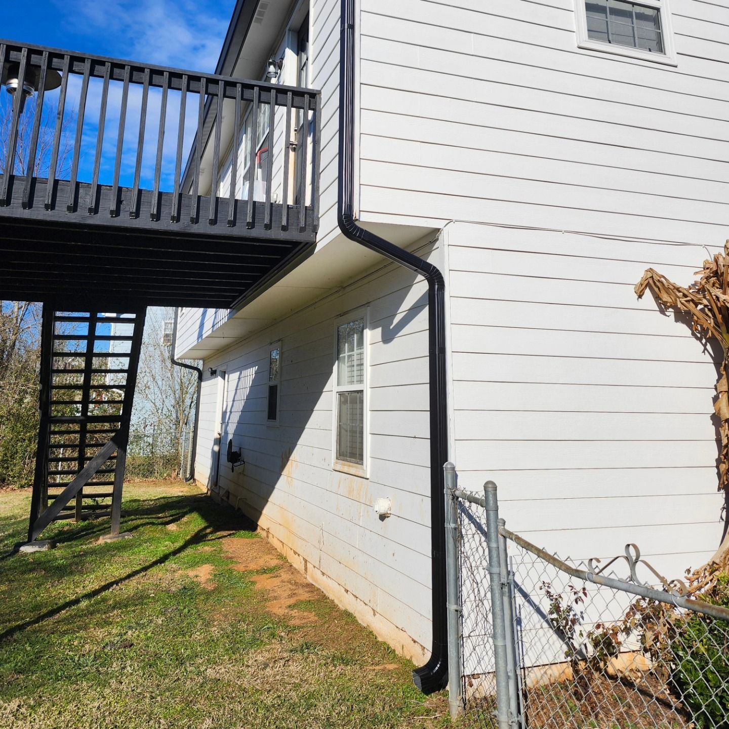 Backyard under a wooden deck beside a white house with a black downspout and chain-link fence