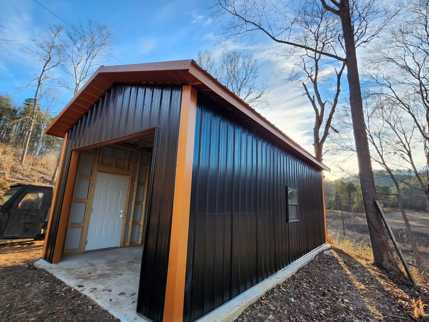 Black metal shed with a white garage door, viewed at an angle in a wooded outdoor setting.