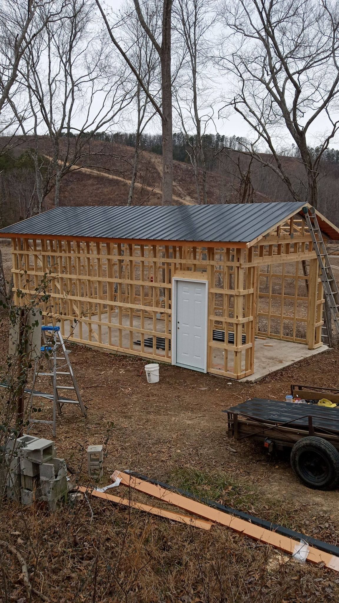 Wood-framed shed under construction with blue metal roof in a wooded yard.