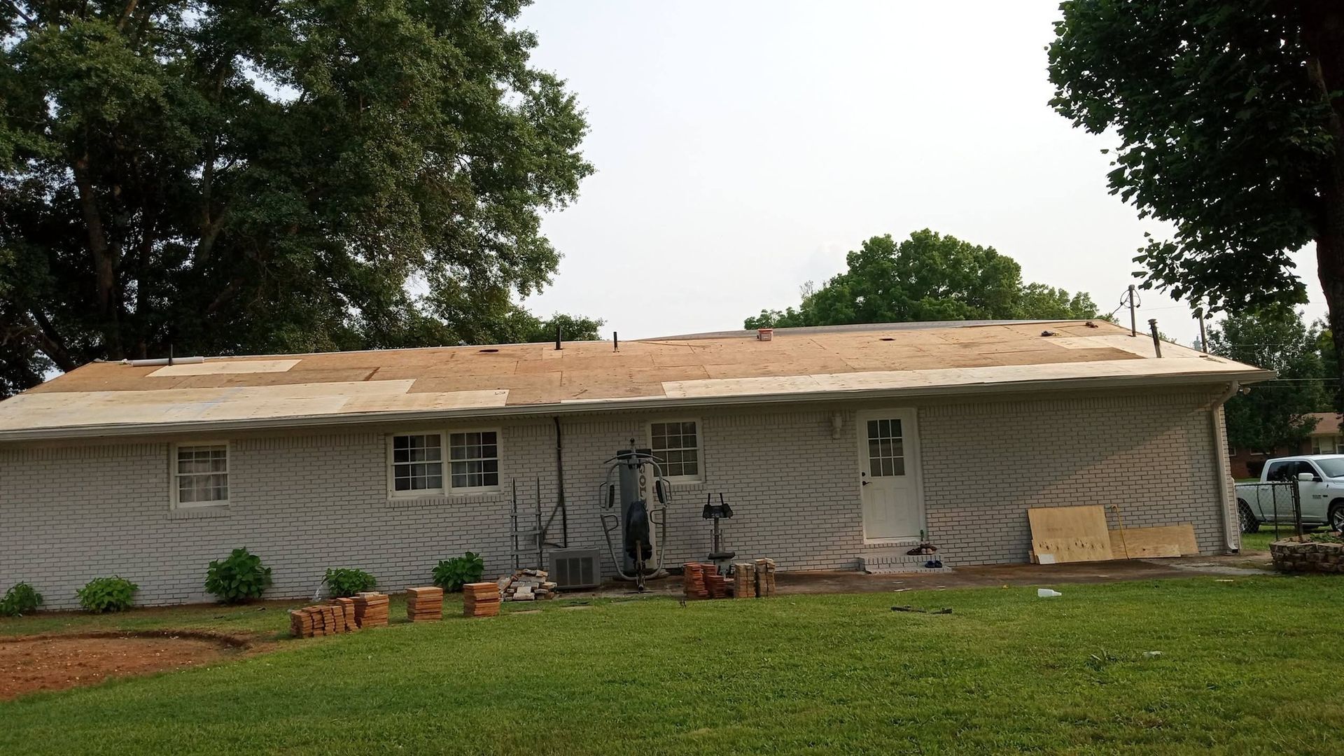 Single-story beige house with a worn roof, front porch, and lawn under cloudy skies
