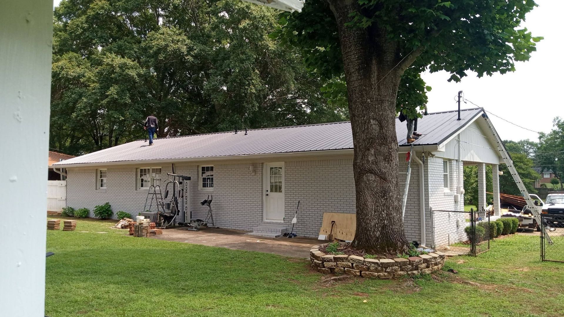 Single-story white house with a metal roof, large tree in front, and grassy yard on a cloudy day