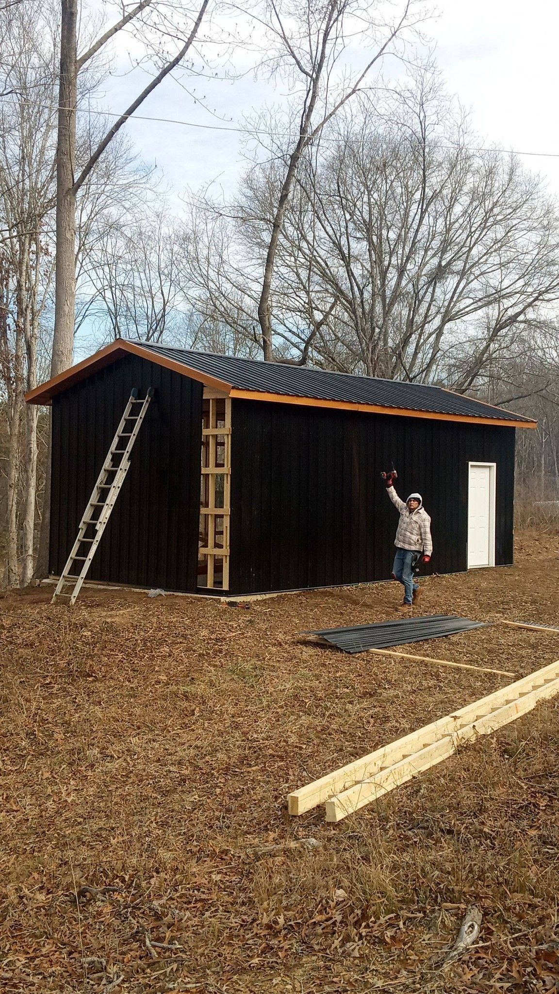 Small black shed under construction in a wooded yard, with a ladder and person standing nearby.