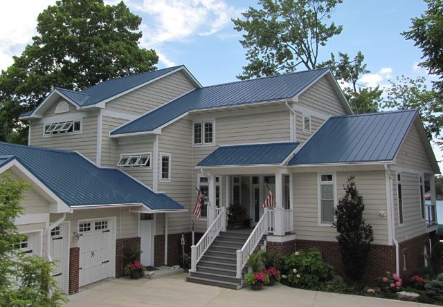 Large beige house with blue metal roofs and front steps, surrounded by trees and flowers