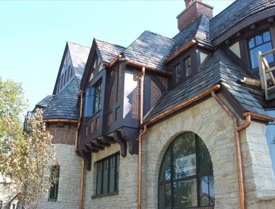 Stone house with steep slate roof, arched windows, and dark timber trim under a clear blue sky