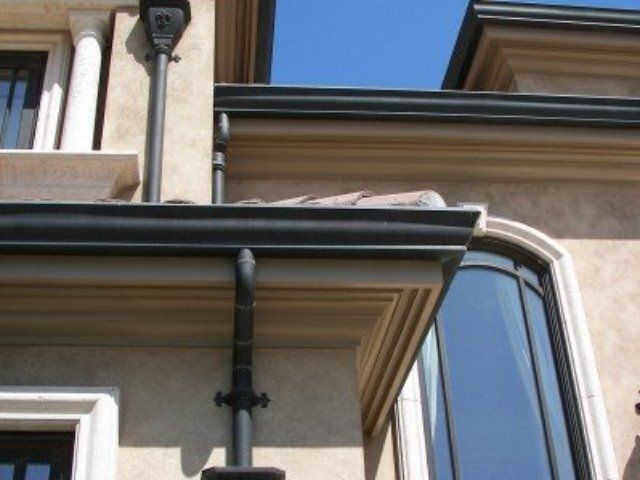 Exterior corner of a stucco building with dark trim, a gutter, and a blue-tinted bay window under a blue sky
