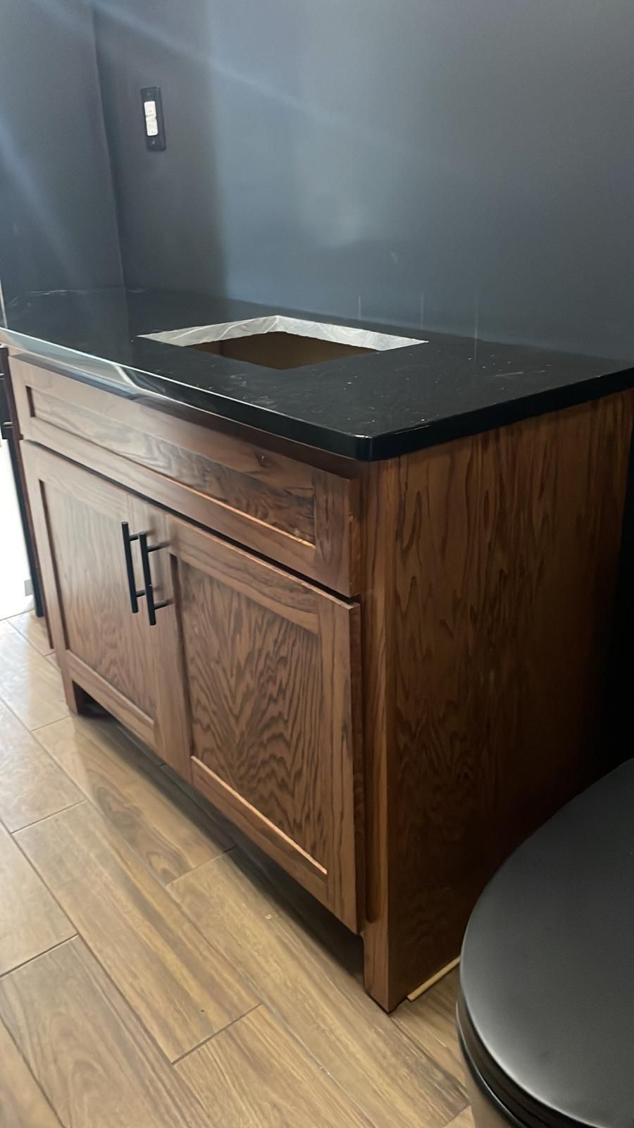 Wooden bathroom vanity with black countertop and sink cutout against a dark wall.