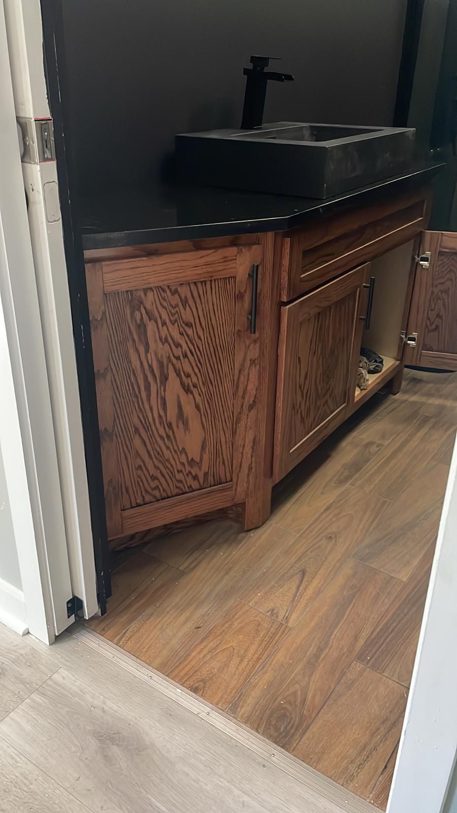 Wooden bathroom vanity with black sink and faucet against a dark wall, viewed from the doorway