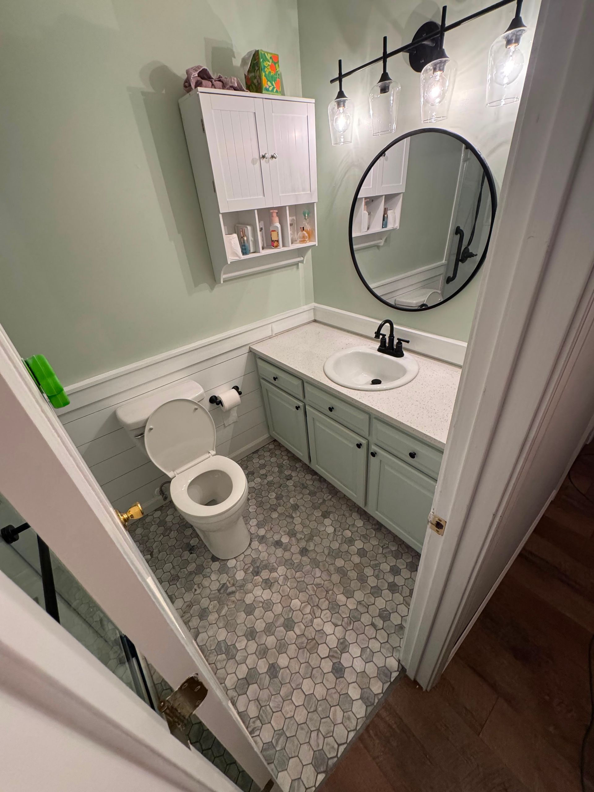 Small bathroom with white walls, green vanity, round mirror, and mosaic tile floor, viewed from the doorway