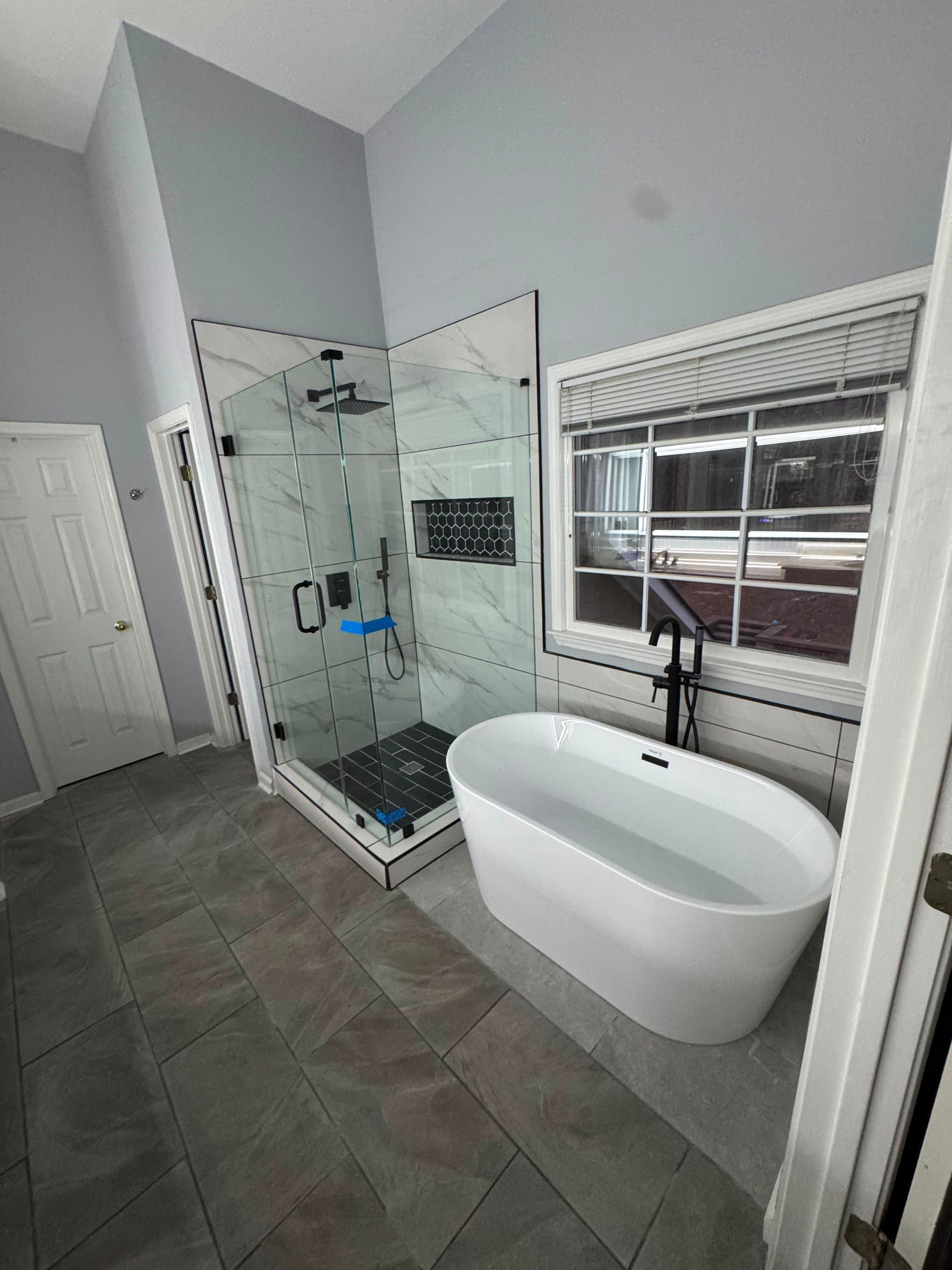Modern bathroom with glass shower, freestanding tub, gray tile floor, and window blinds
