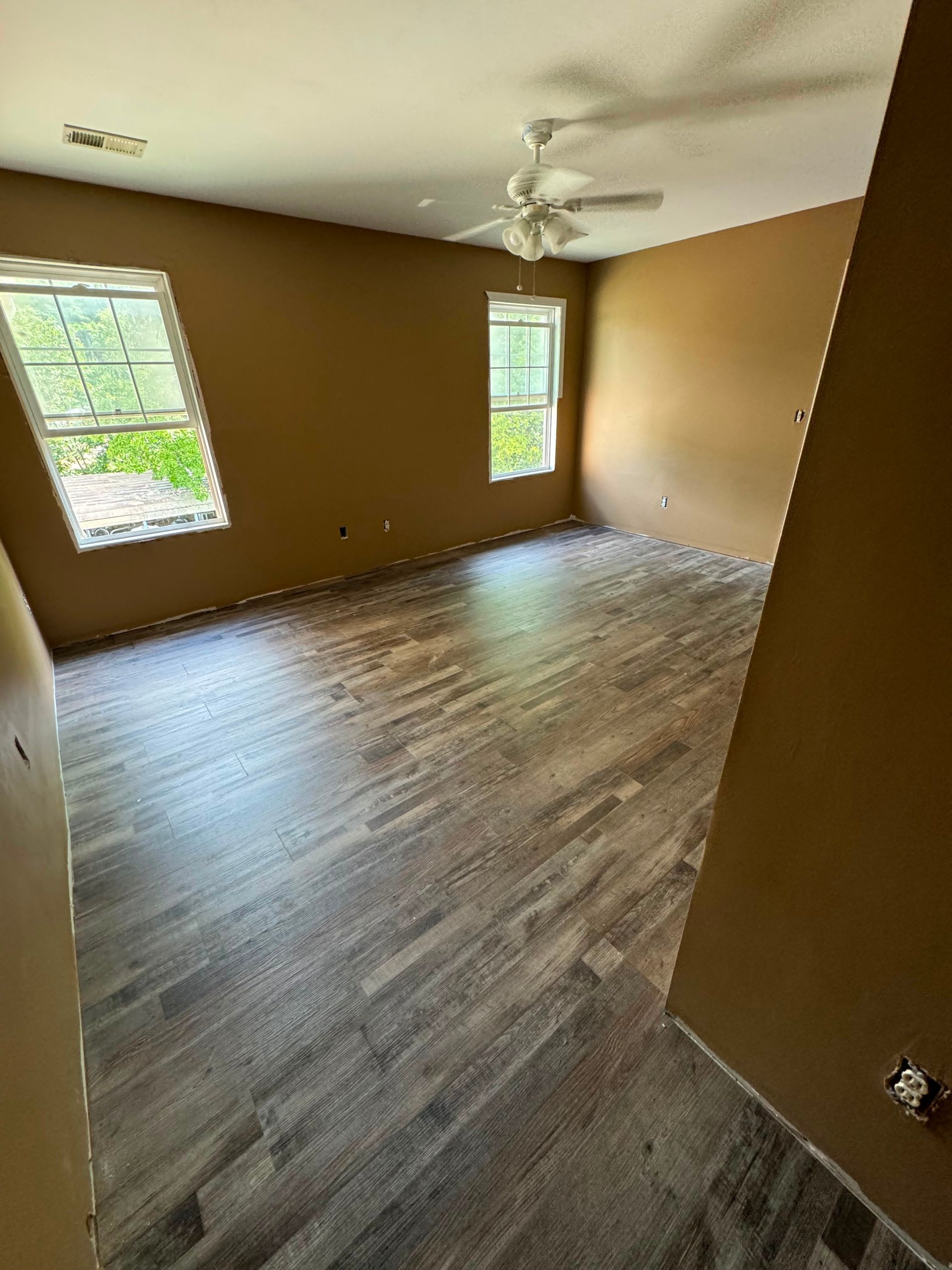 Empty room with brown walls, wood-look flooring, white ceiling fan, and two windows.