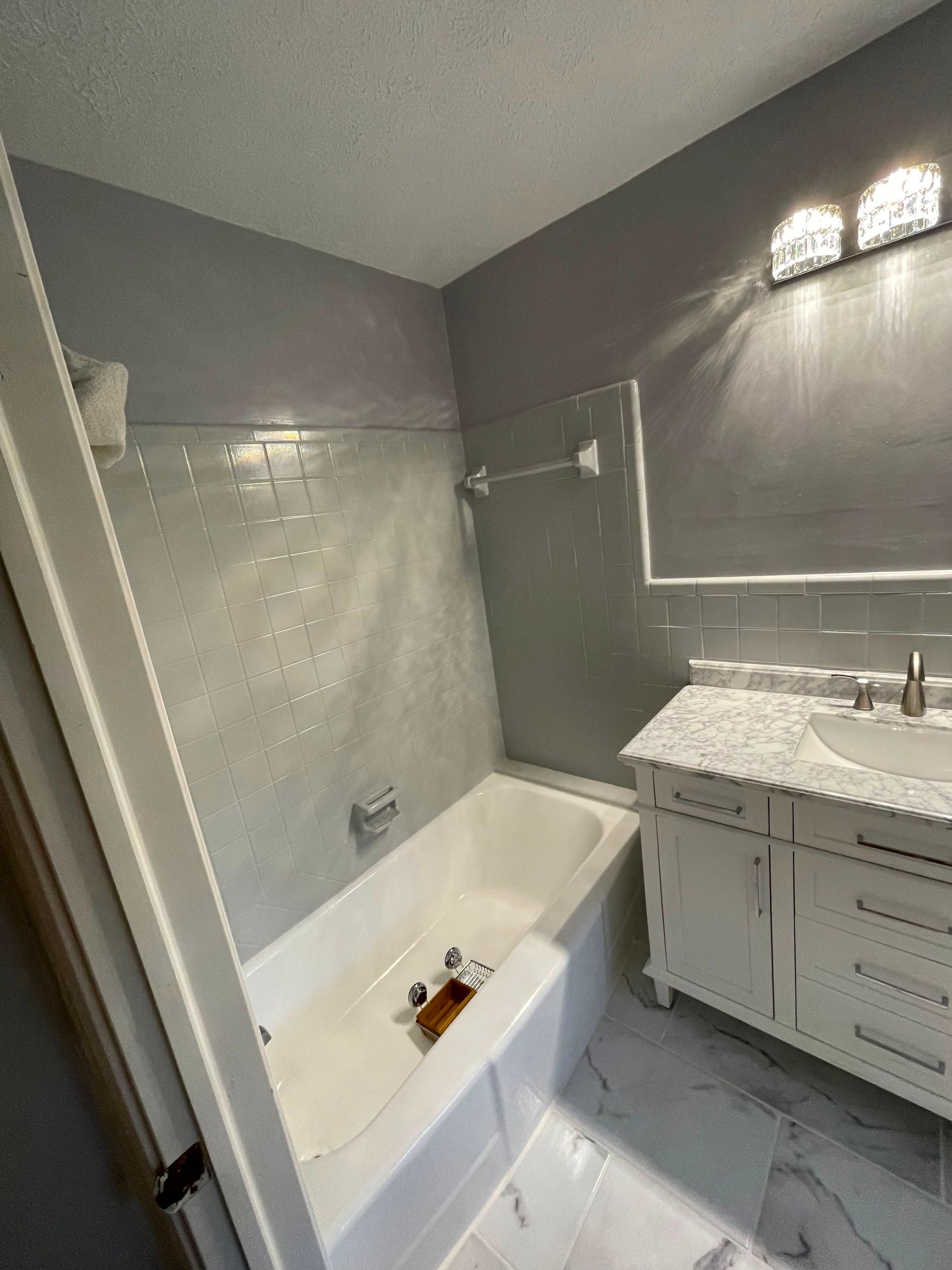 Small bathroom with white tub, gray tiled walls, and a white vanity sink under lights.