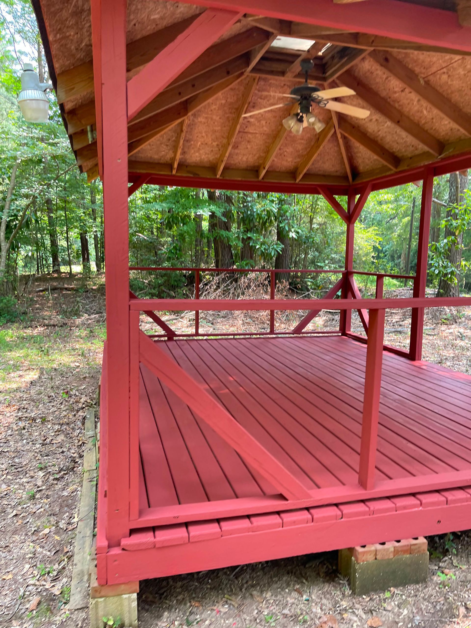 Red-painted wooden gazebo with railing and ceiling fan in a wooded yard