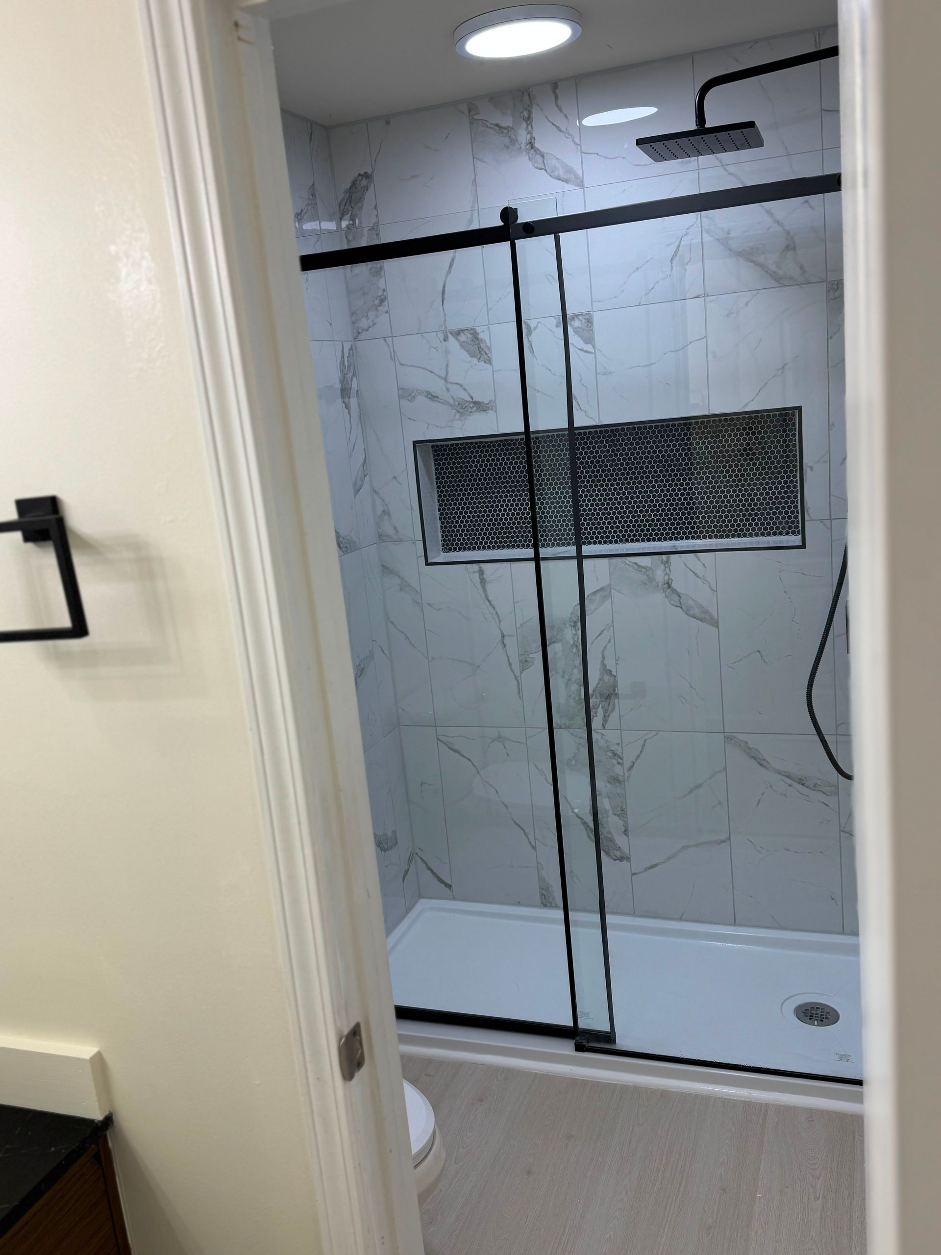Bathroom with glass shower enclosure, marble-look tile, and black fixtures viewed through an open door