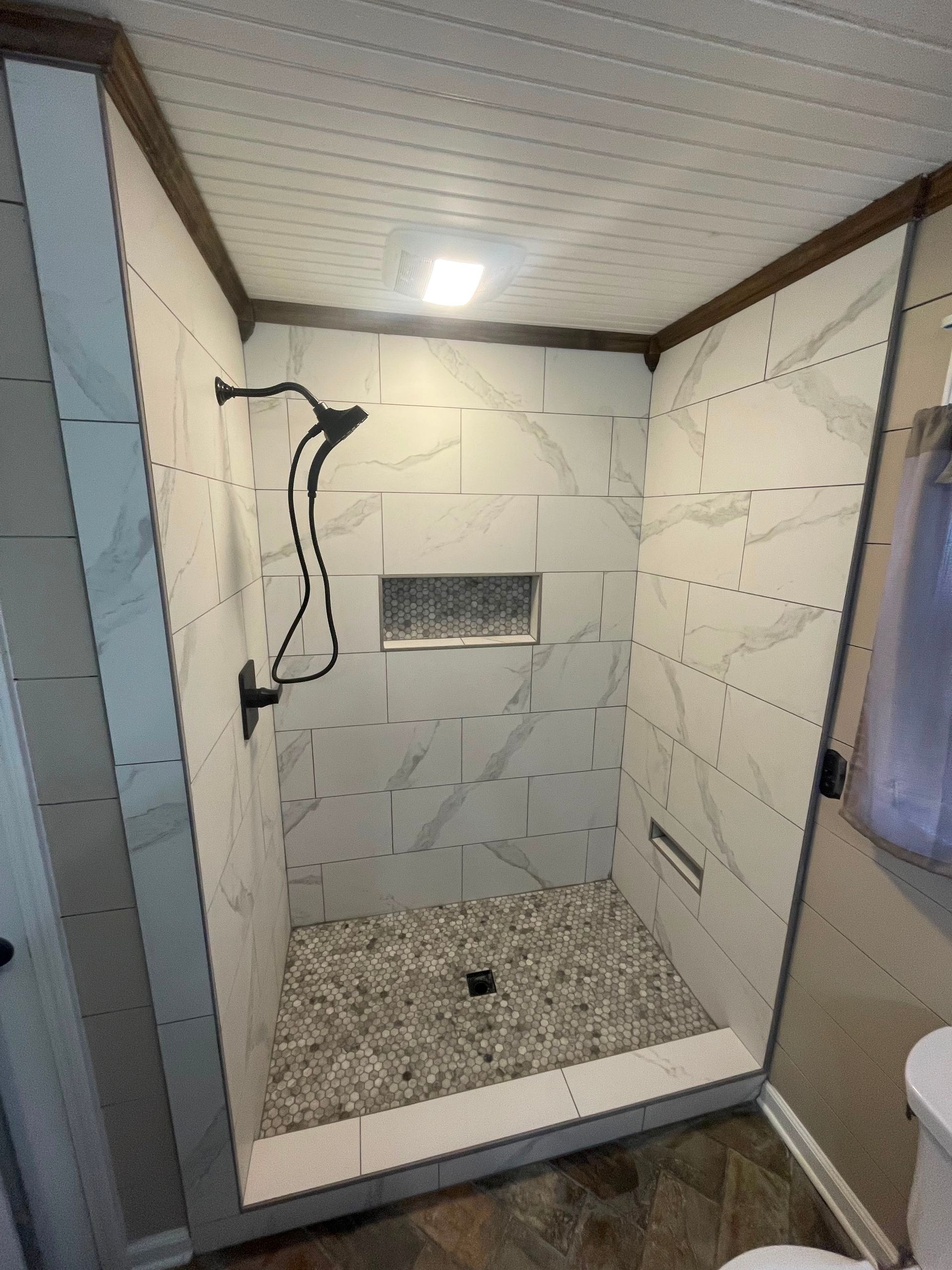 Small tiled walk-in shower with white walls, pebble floor, and a recessed shelf under bright ceiling light