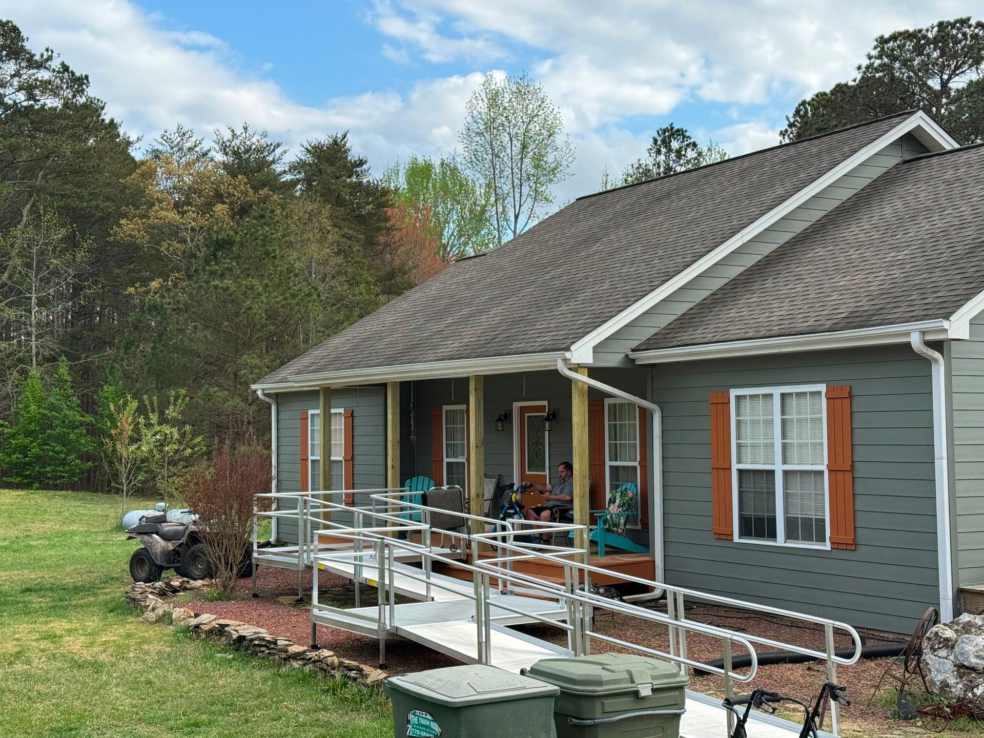 Gray house with a front porch ramp and railings, set beside a grassy yard and trees under a blue sky.