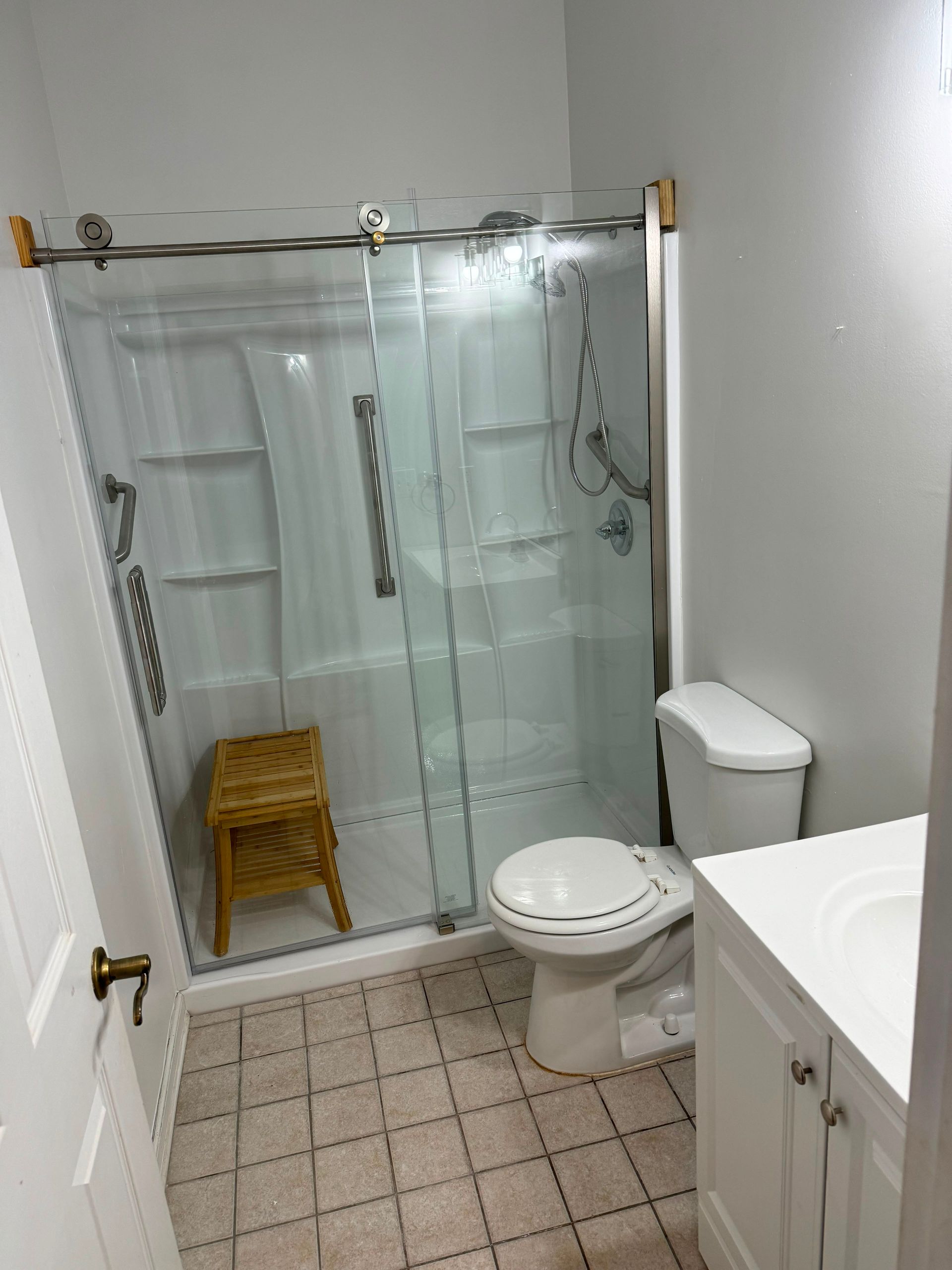 Small bathroom with glass shower, toilet, vanity, and wicker stool on tiled floor.