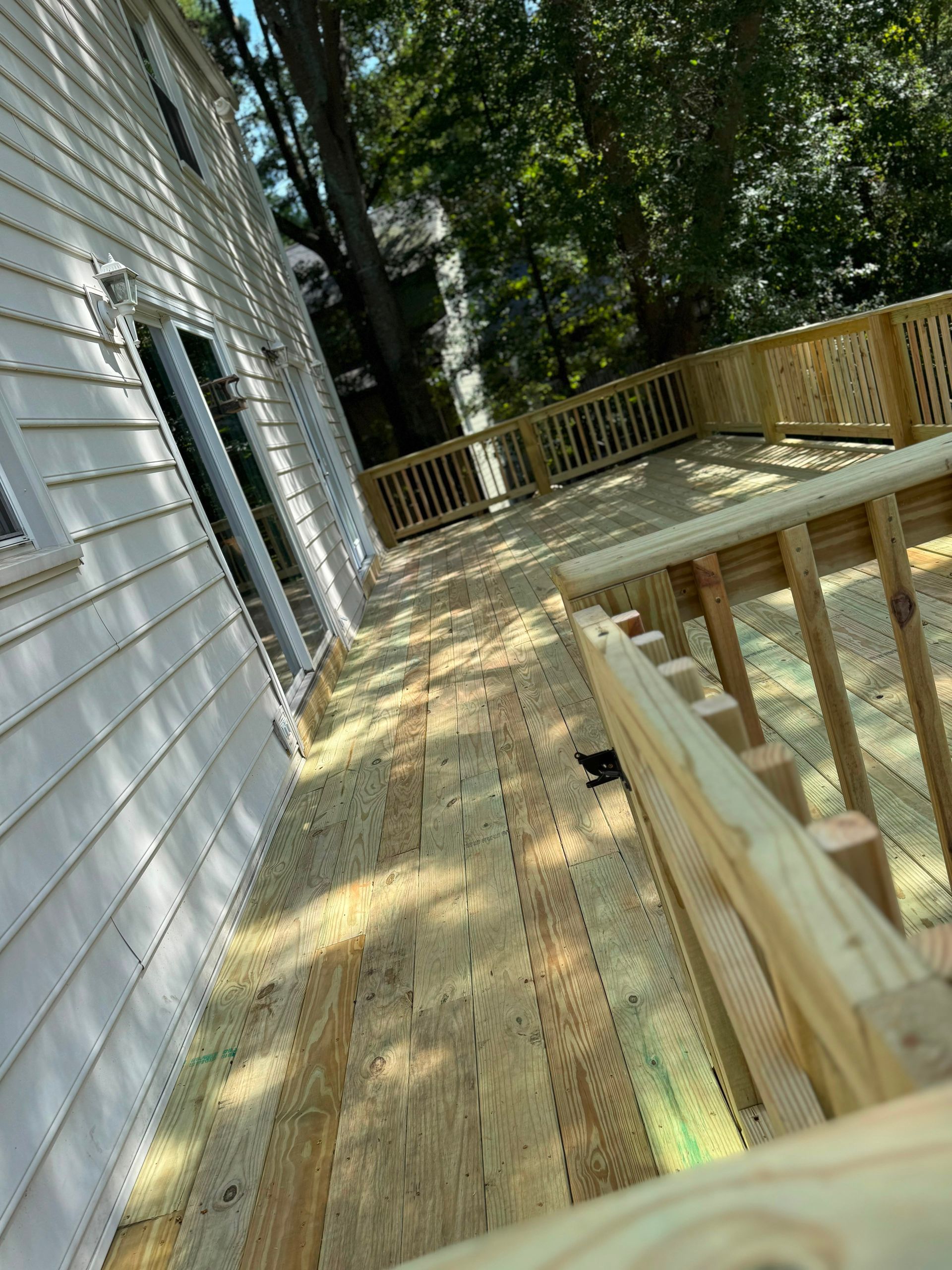 Wooden deck walkway beside a white house, with railings and trees overhead in dappled sunlight.