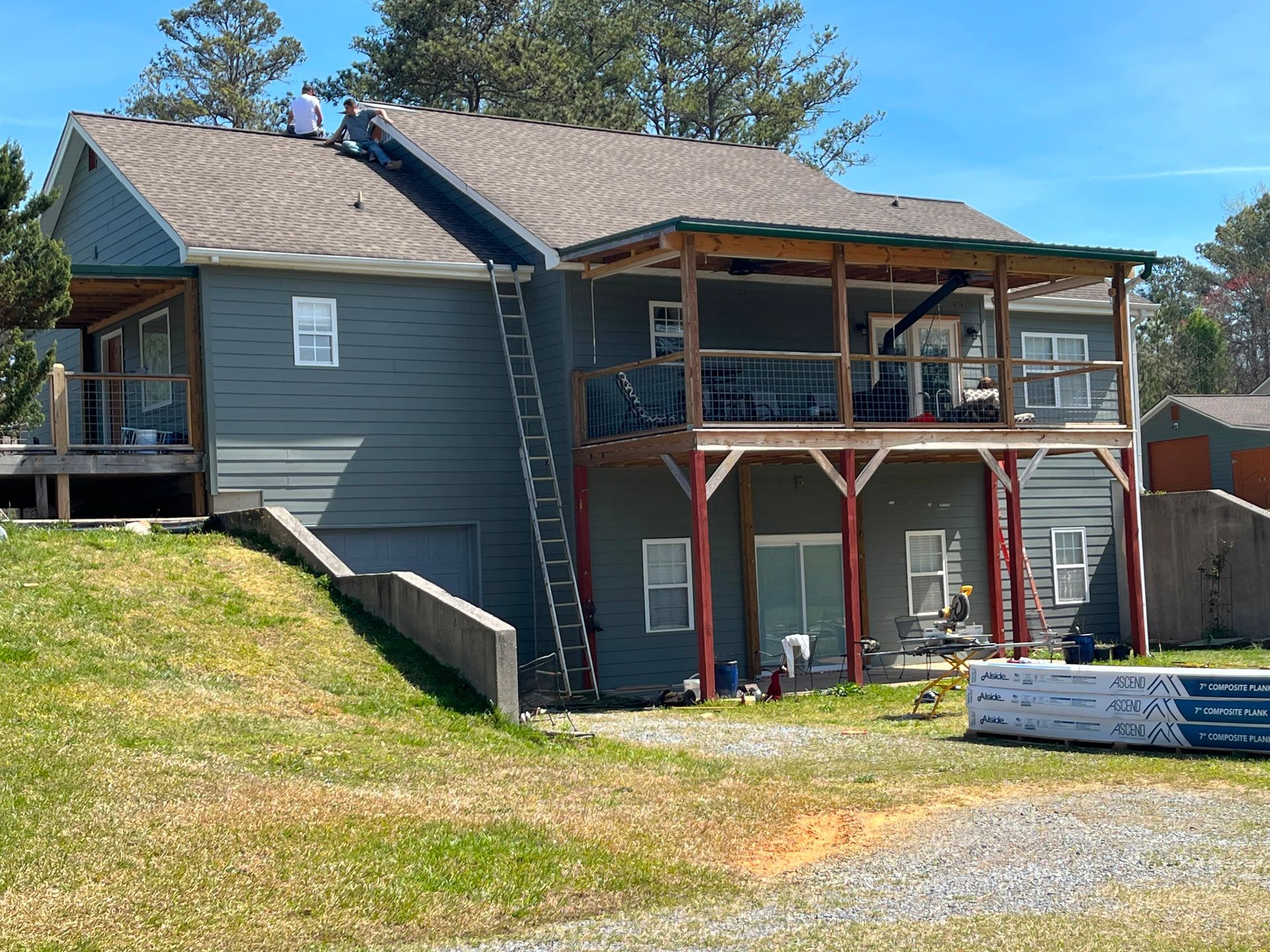 Two-story gray house with deck and ladder on a grassy hillside, viewed from a gravel driveway