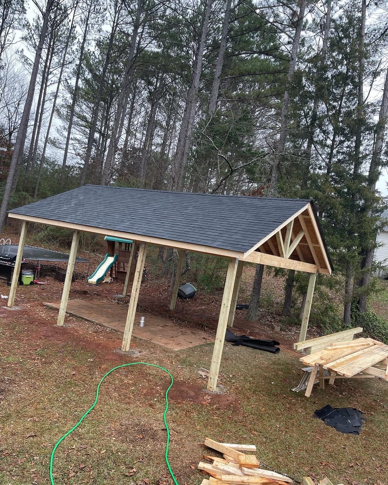 Partially built wooden shelter with dark roof in a wooded yard, construction materials scattered nearby.