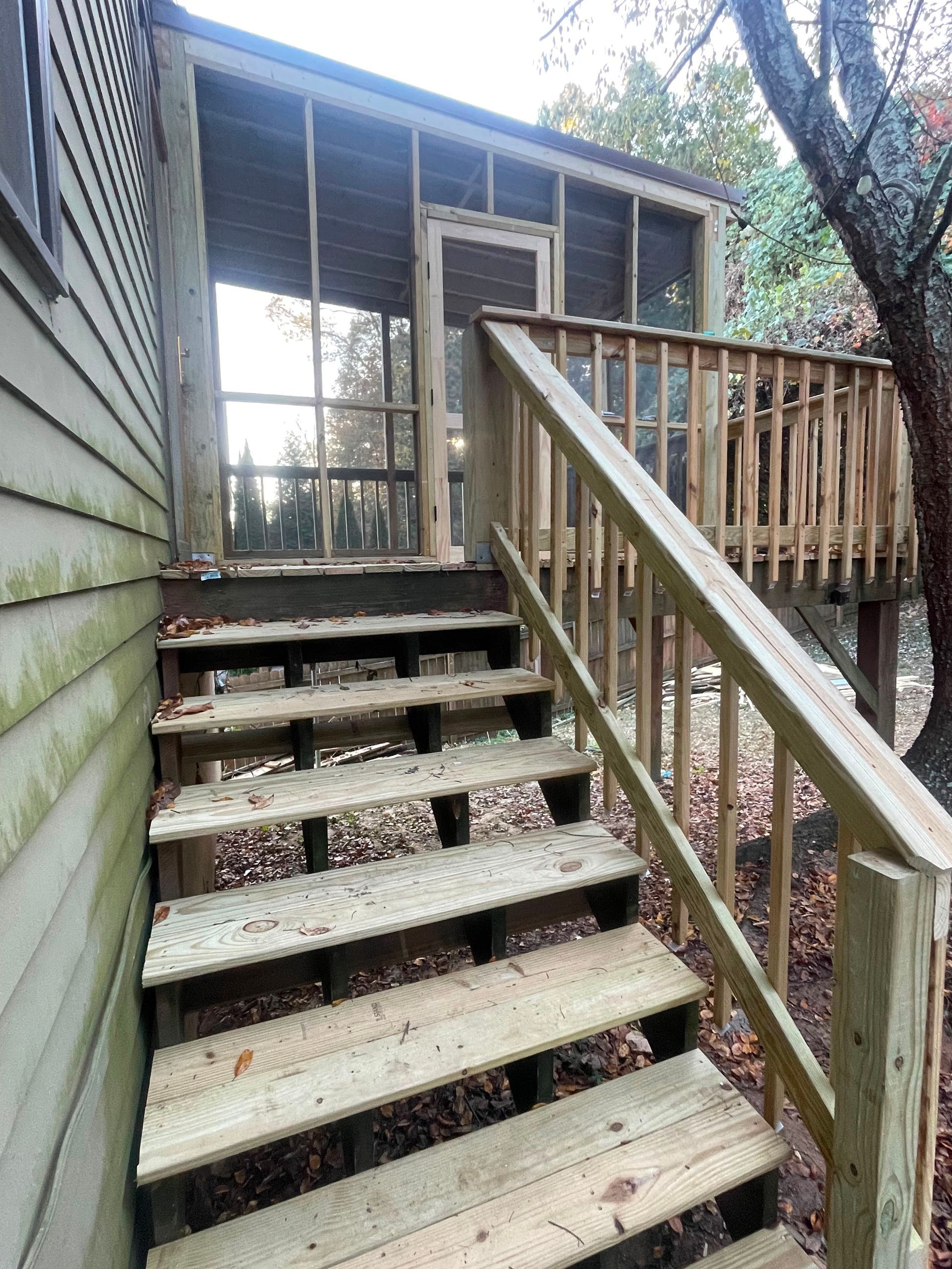 Weathered wooden stairs lead up to a screened porch beside a house, with a handrail and trees around it.