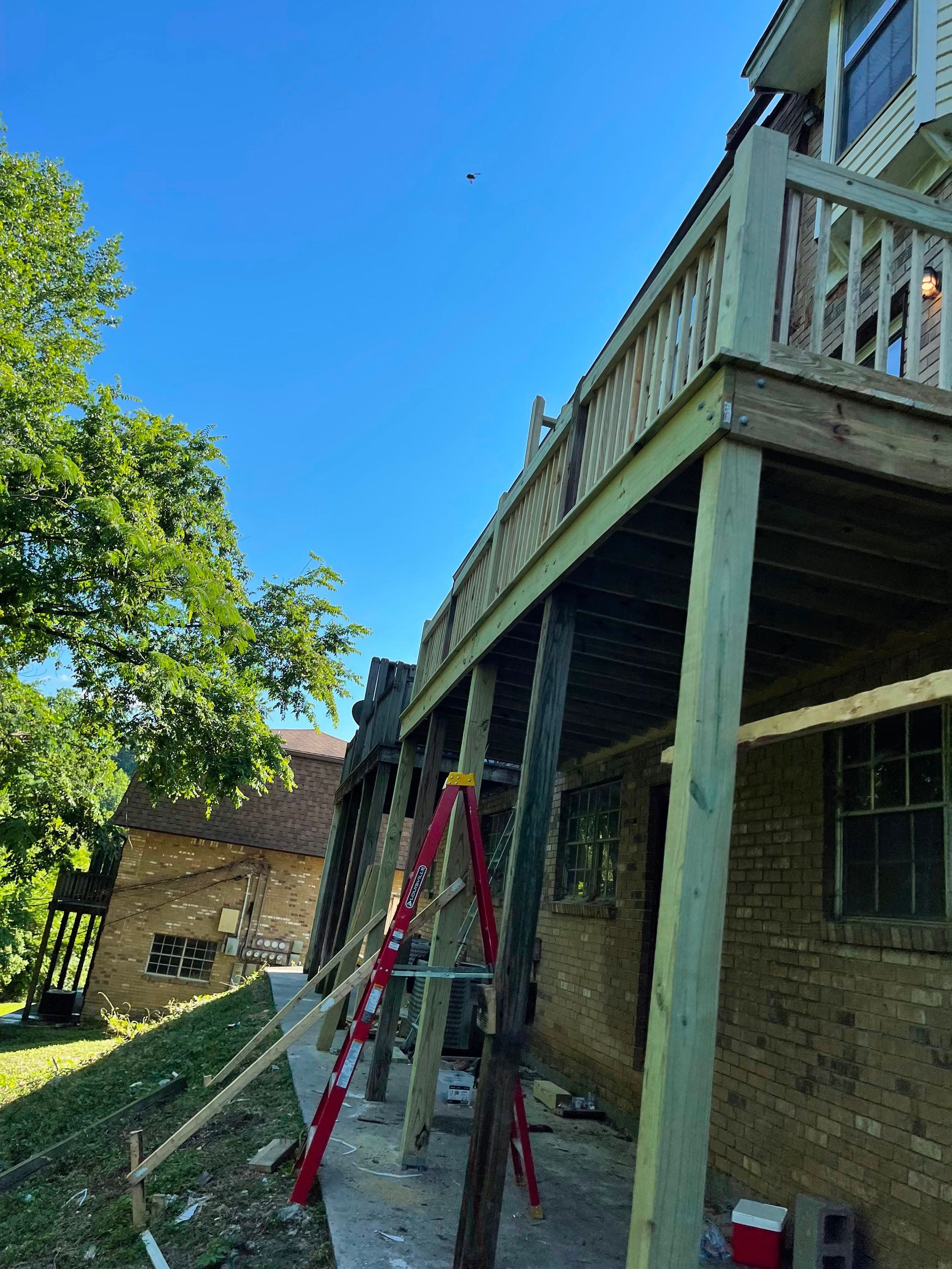 Backyard deck under repair with red ladder, wooden posts, and brick house in sunlight