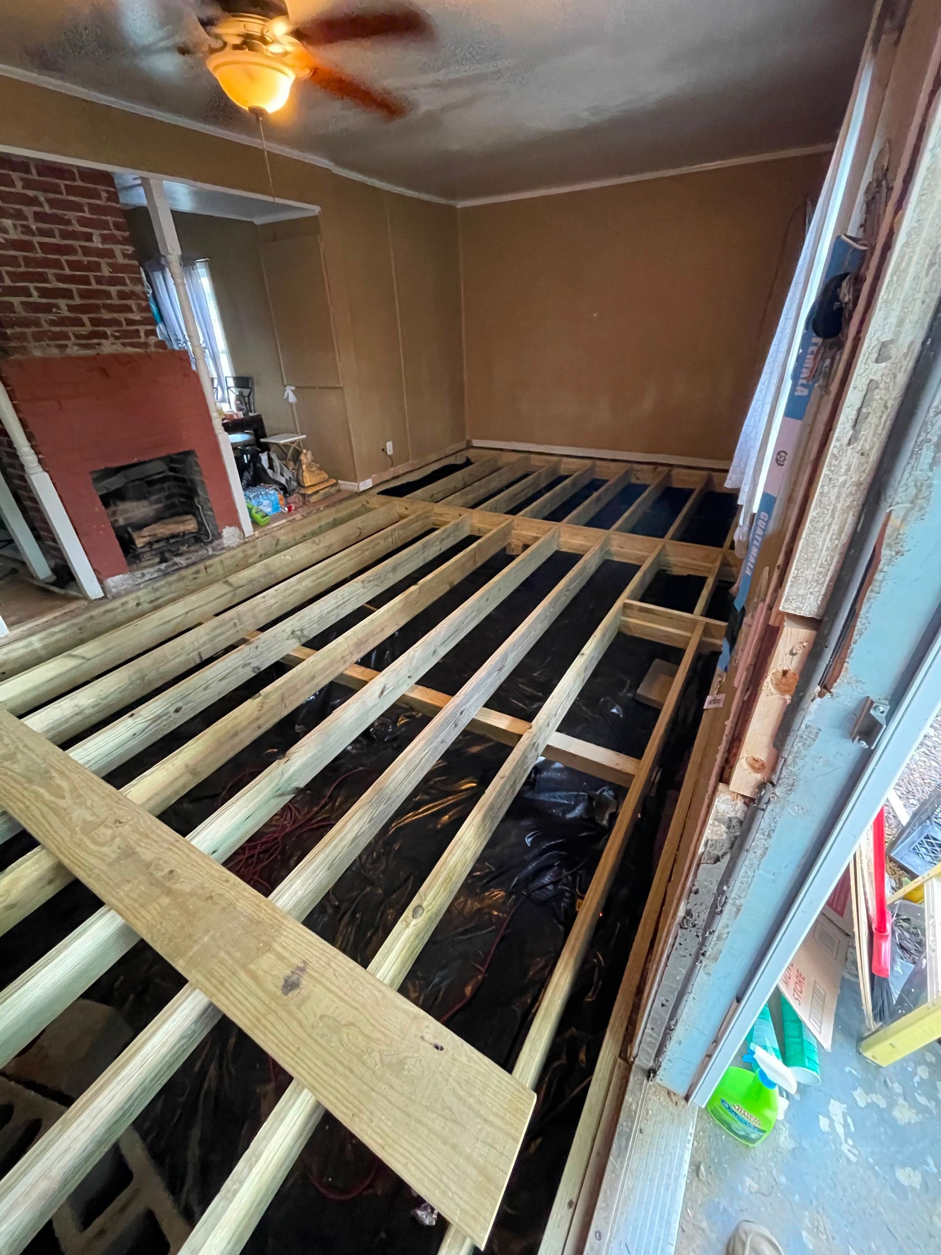 Room under renovation with exposed floor joists, fireplace, and ceiling fan.