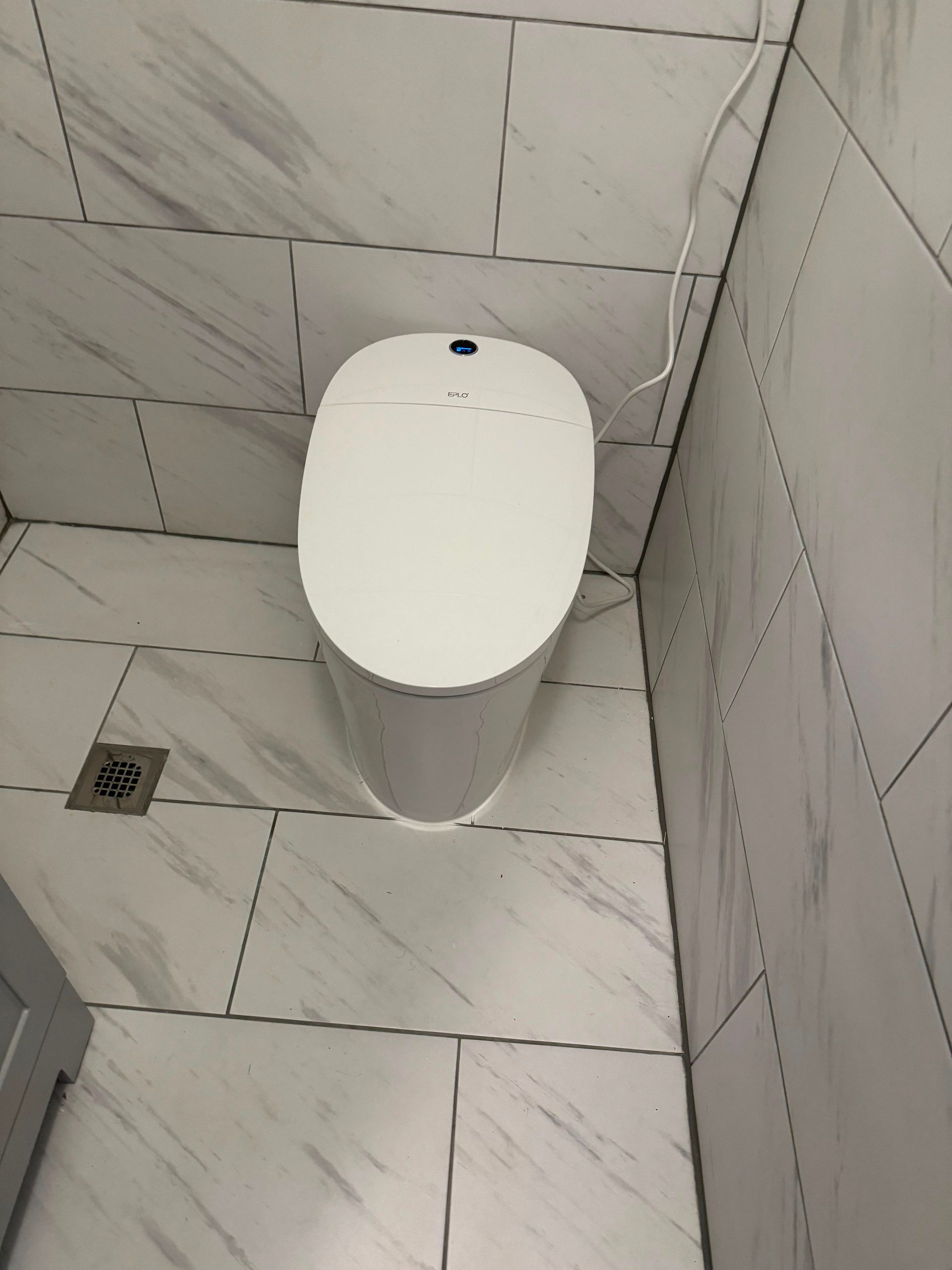 White wall-mounted toilet in a tiled bathroom corner with a floor drain nearby