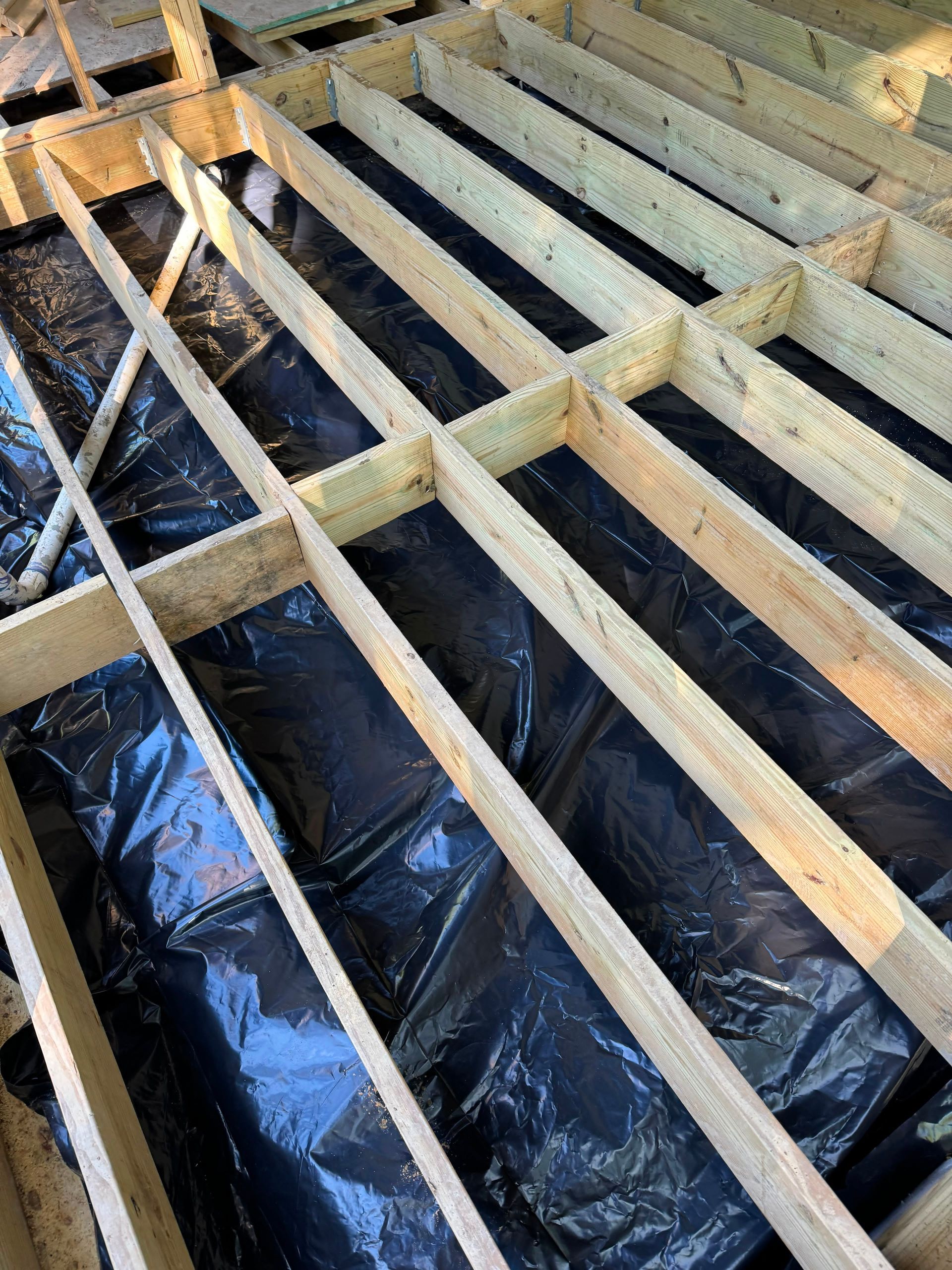 Exposed wooden floor joists over black plastic vapor barrier in an unfinished room