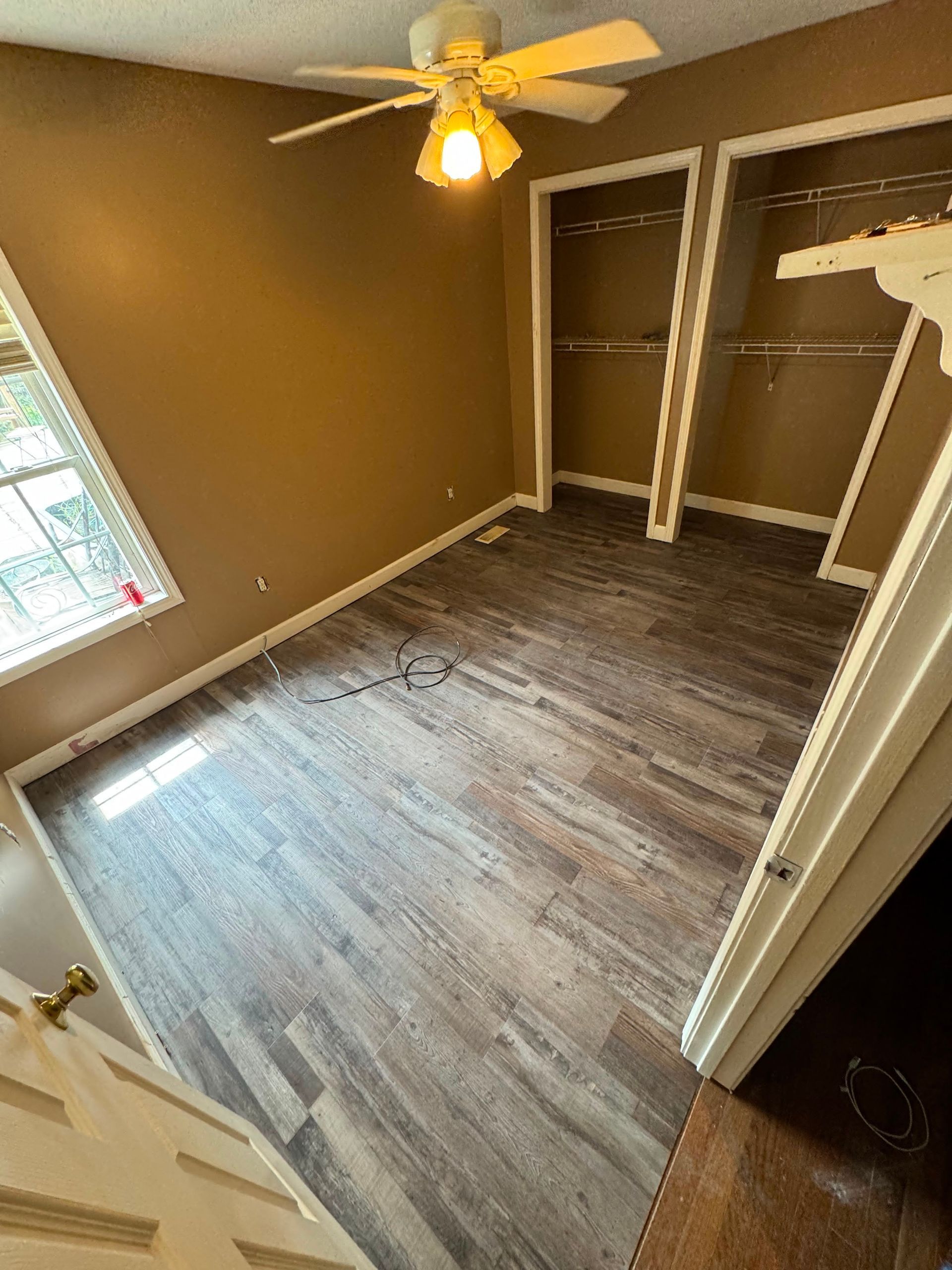 Empty bedroom with brown walls, gray wood-look floor, ceiling fan, closet, and a window.