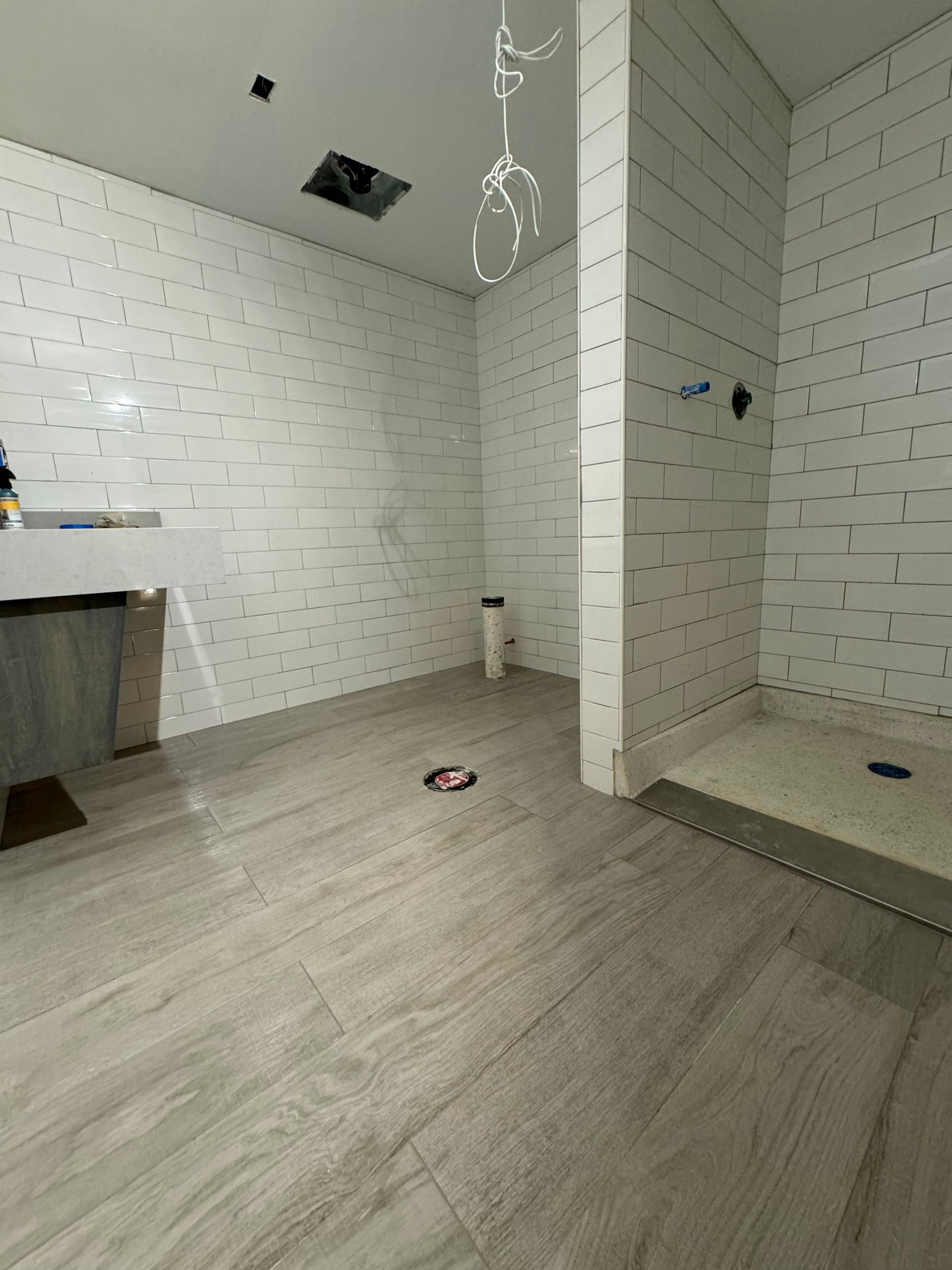 Empty tiled bathroom under renovation with shower area, hanging wires, and light wood-look floor.