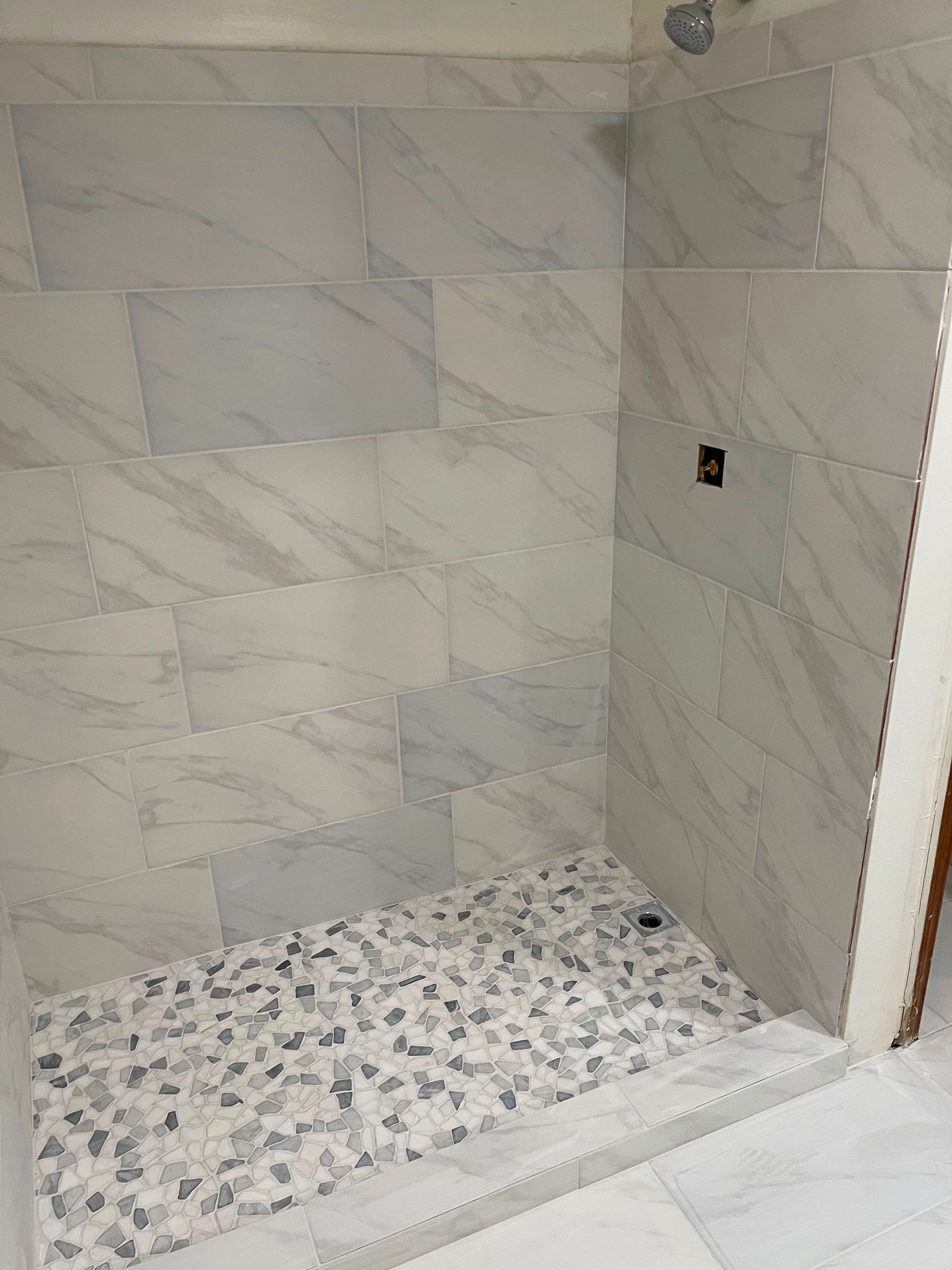 Tiled shower alcove with marble-look walls and a pebble mosaic floor, unfinished at the right edge