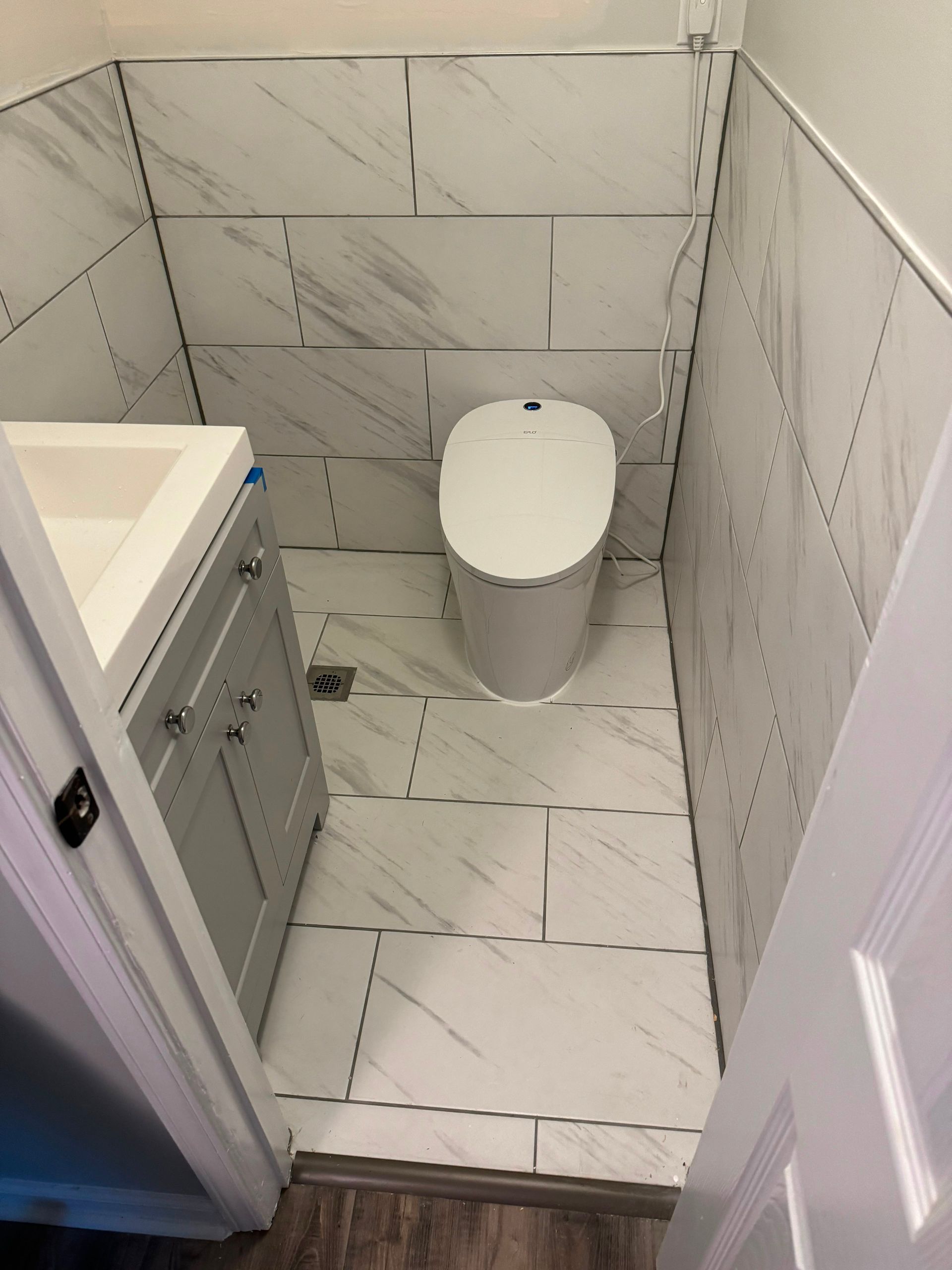 Small tiled bathroom with a toilet and vanity sink, viewed through an open door.