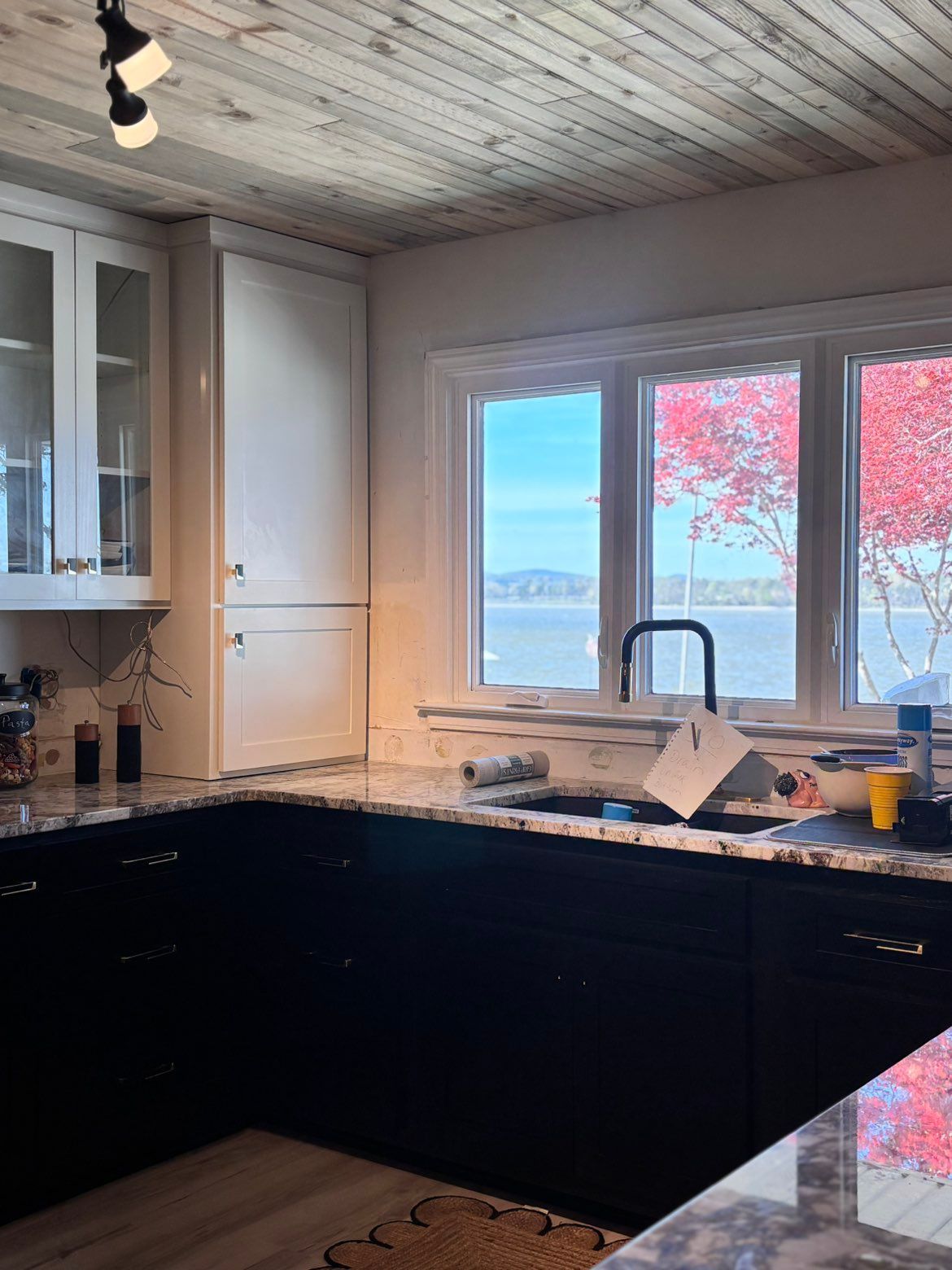 Modern kitchen with dark cabinets, white walls, and large windows overlooking water and red autumn trees