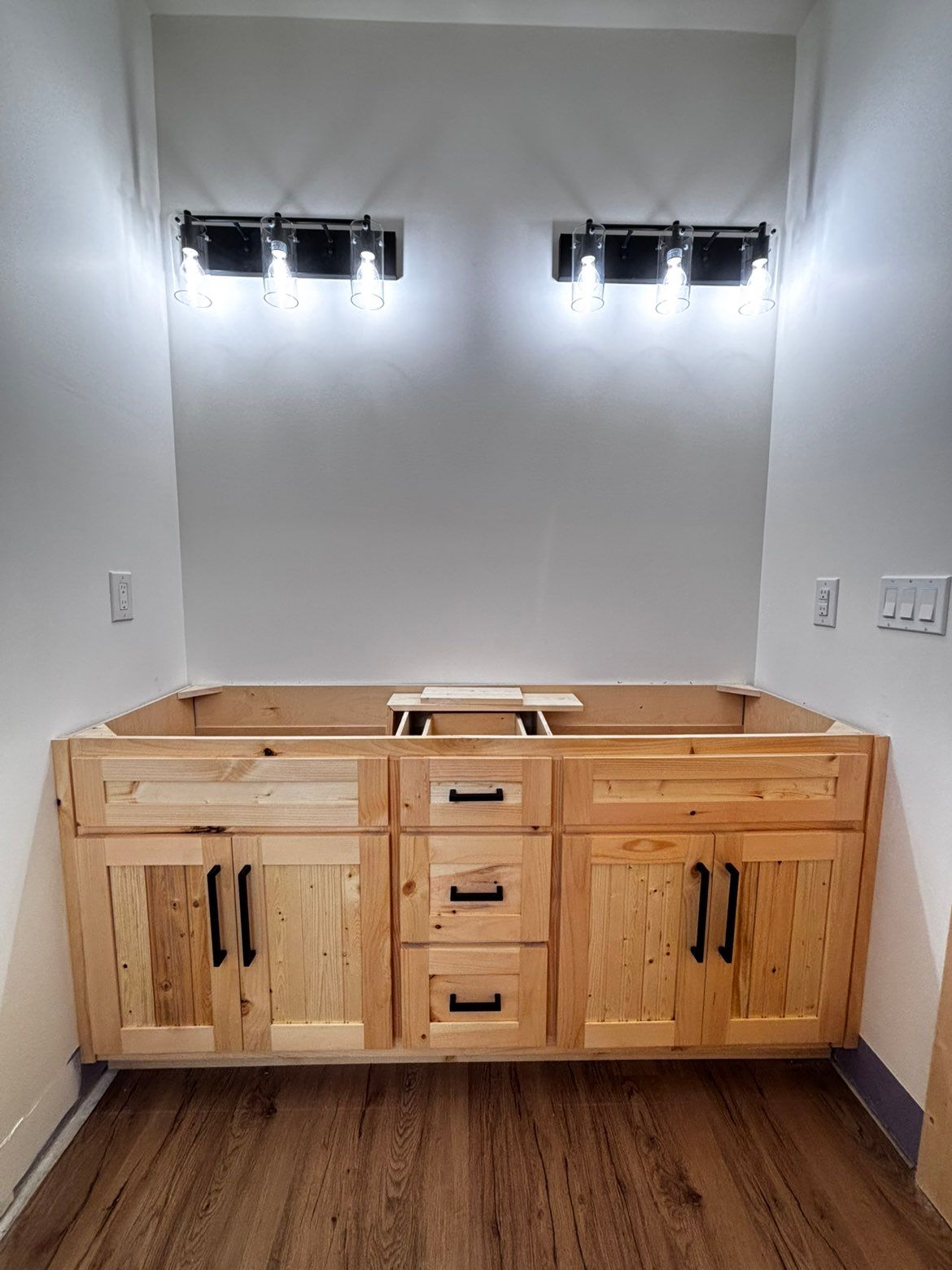 Unfinished bathroom vanity with wood cabinets and black fixtures beneath two wall lights.